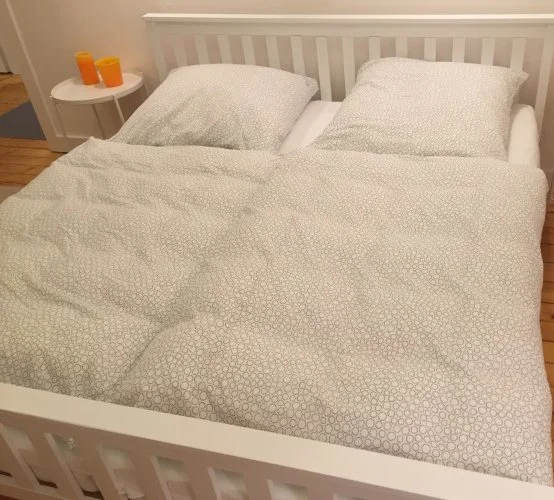 Ein Bett mit weißen Bettwäsche und zwei Kissen, daneben ein kleiner runder Nachttisch mit zwei orangefarbenen Kerzen. Das Bett befindet sich in einem Schlafzimmer mit Holzfußboden.