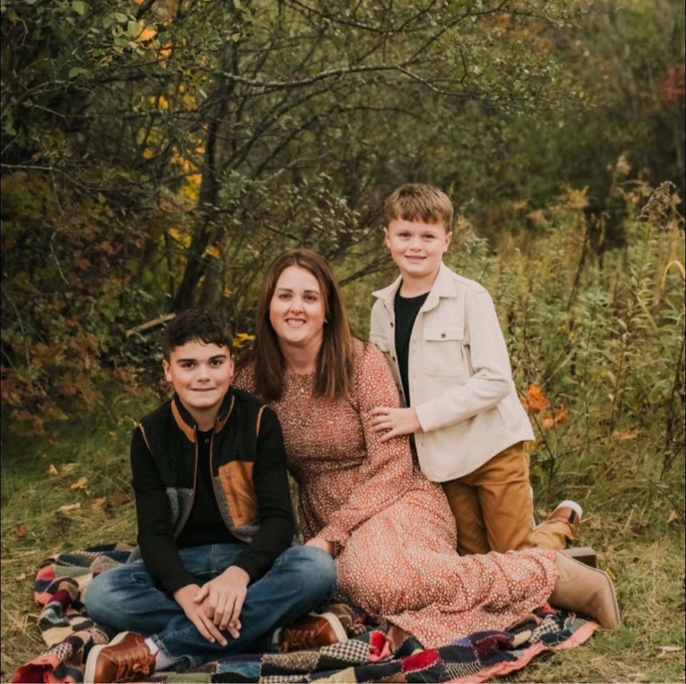 Mother and two young sons sitting on a blanket in a wooded area during fall.