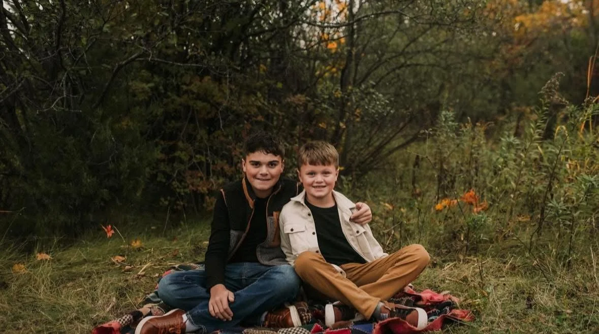 Two boys sitting on a blanket outdoors in a wooded area during fall, smiling at the camera.