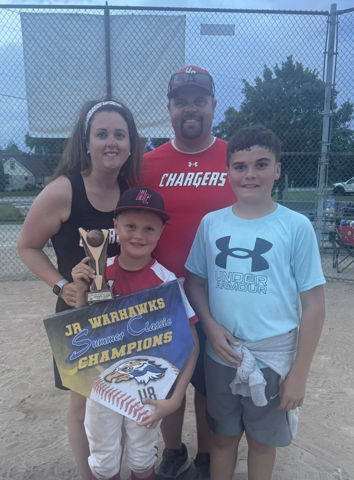 A family of four at a baseball field. The young boy in the front is holding a trophy and a banner that reads "Jr WARHAWKS Summer Classic Champions." The family appears happy, celebrating their victory.