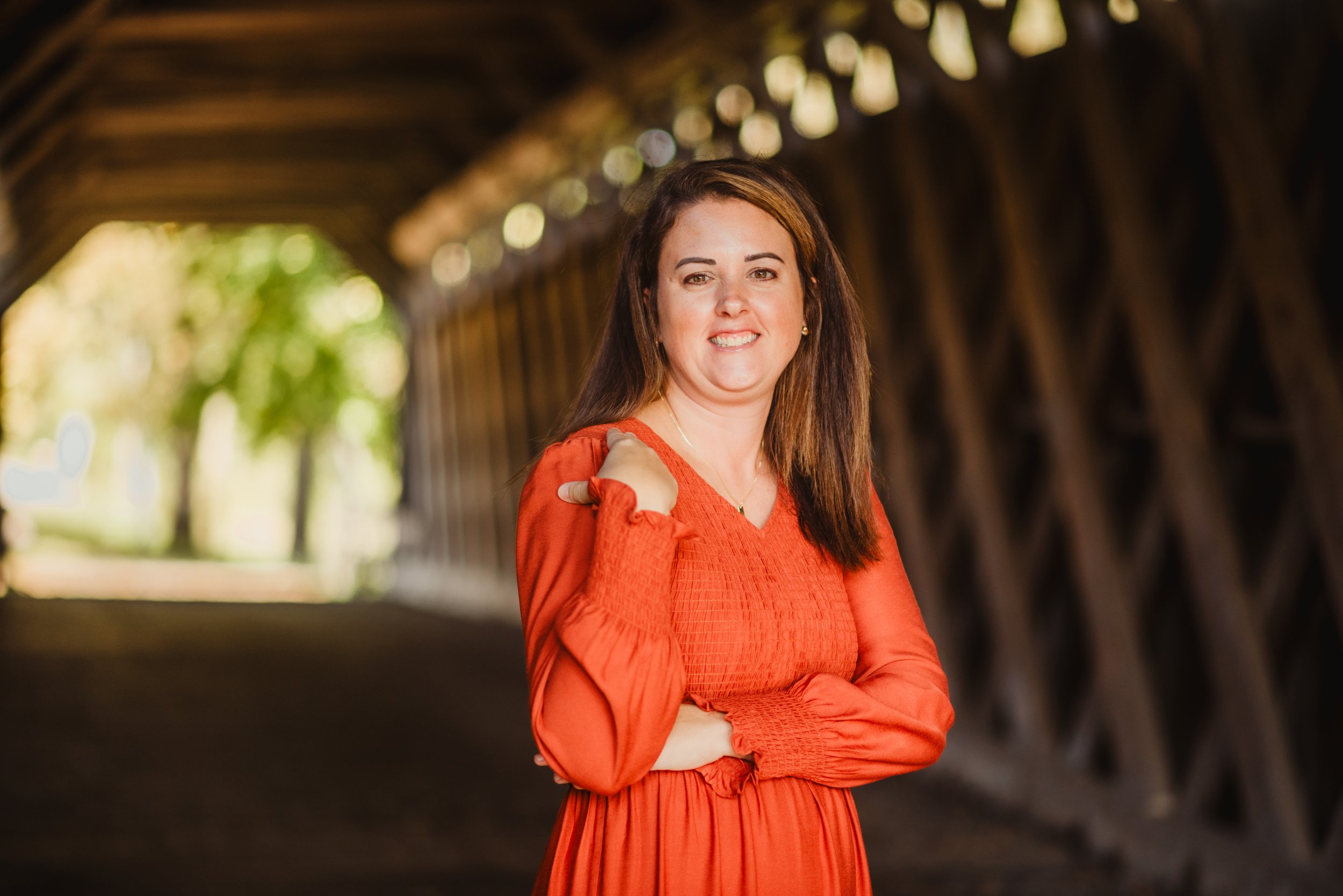 A woman in an orange dress smiling and posing with her left hand on her shoulder inside a wooden tunnel with blurred background of trees and sunlight.