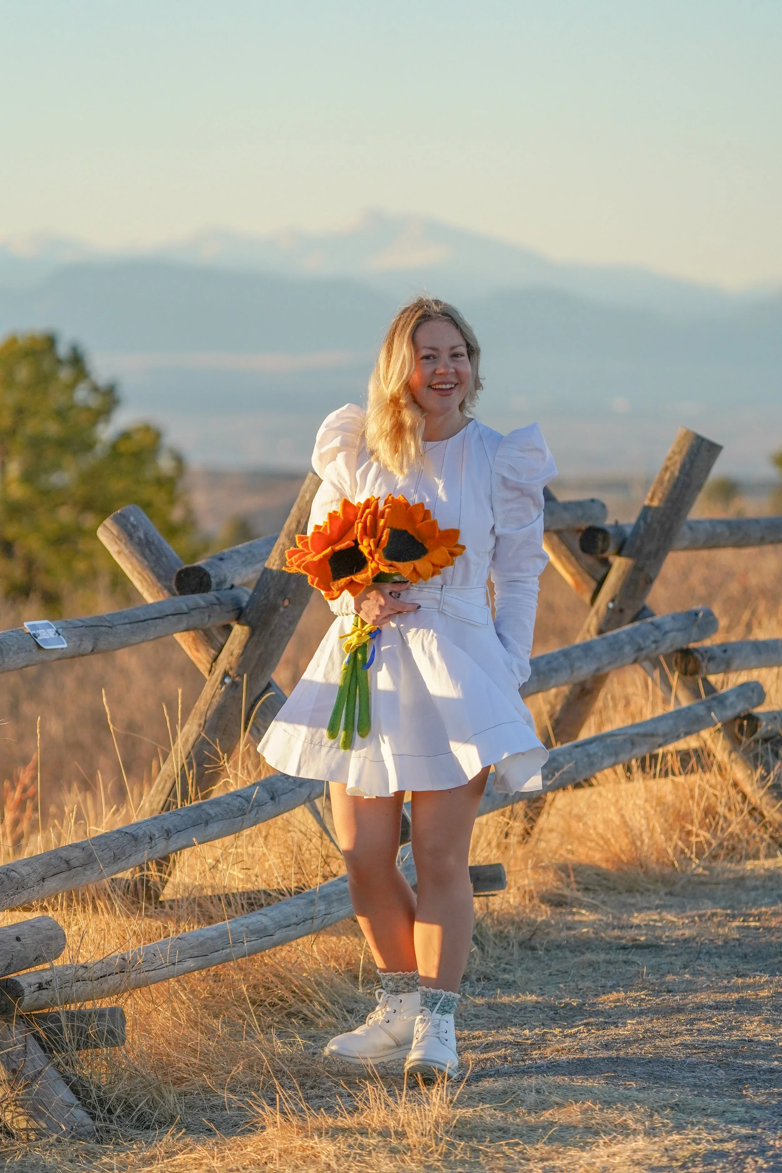 A young woman in a white dress standing outdoors holding a bouquet of sunflowers, with a wooden fence and a mountain in the background during sunset.