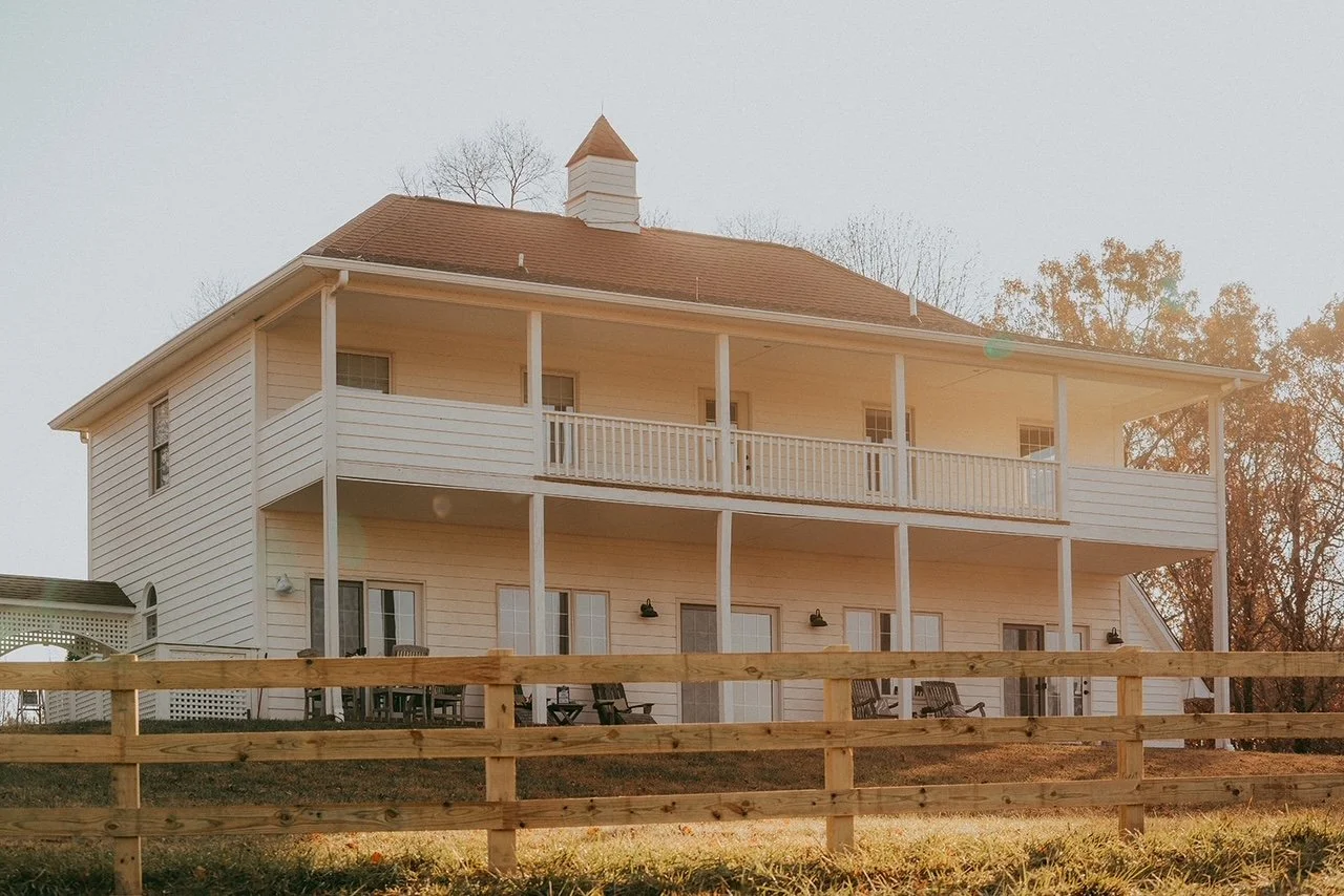 A two-story white house with a brown roof, multiple doors and windows, and a large balcony on the second floor. There is a wooden fence in the foreground, and outdoor furniture and chairs on the ground level patio.