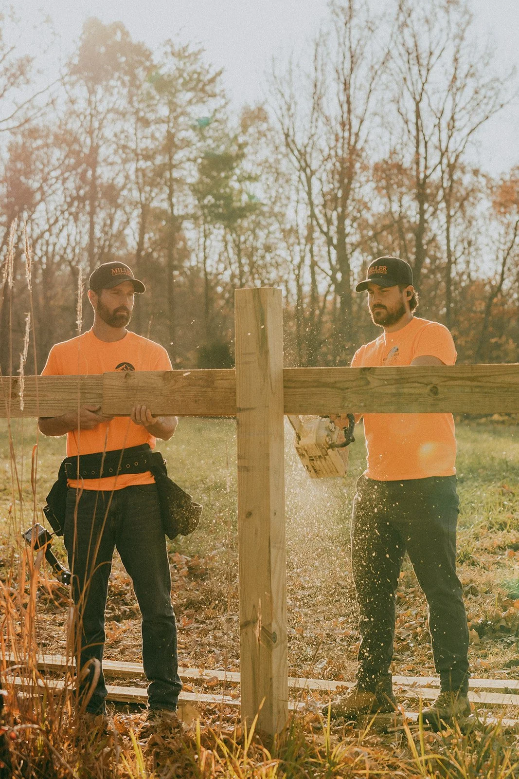 Two men in orange shirts and black caps working outdoors, assembling a wooden structure during late afternoon with trees in the background.