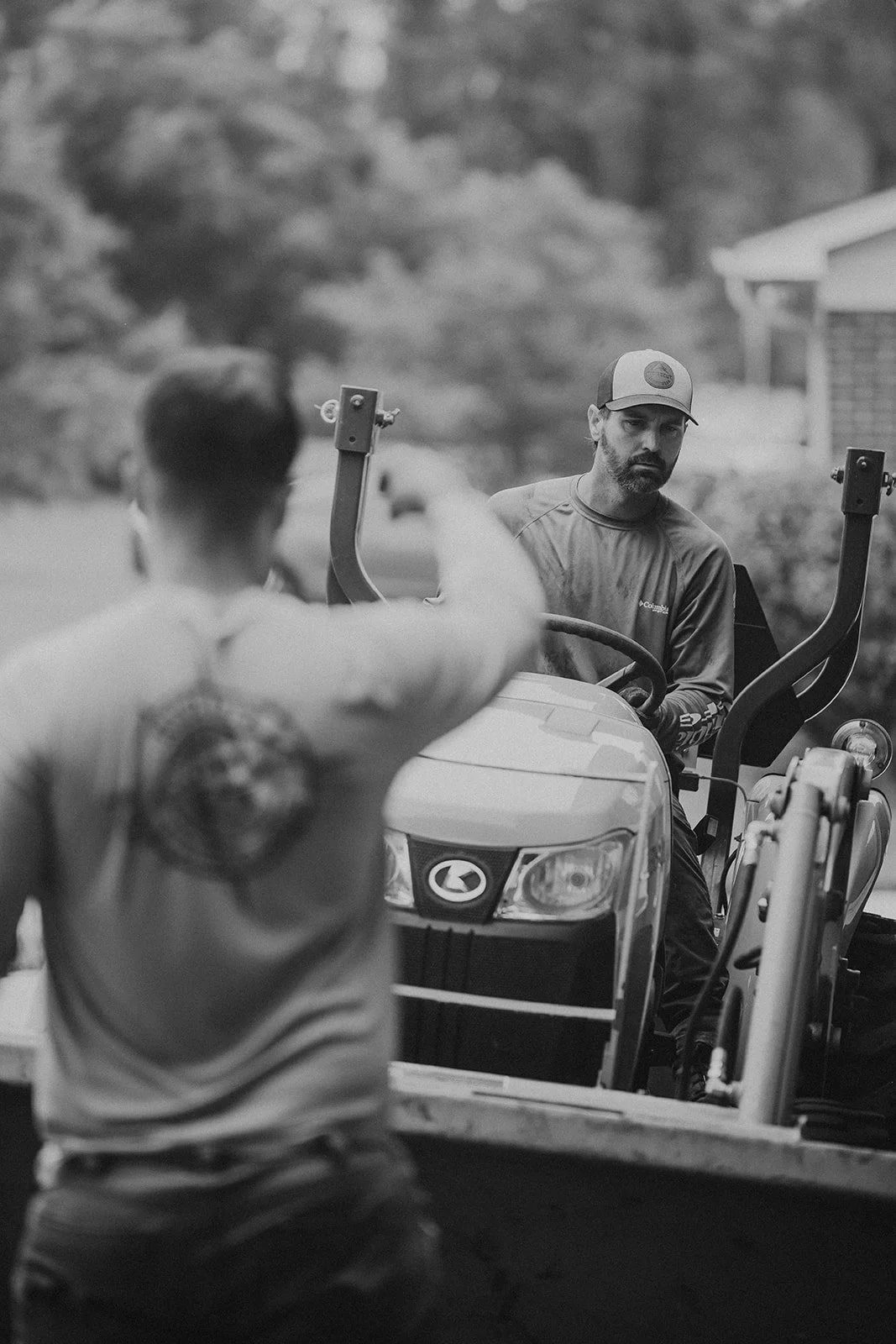 A man with a beard and baseball cap sitting on a tractor, looking at another person with their back to the camera, pointing their finger. The background is outdoors with trees and a house.