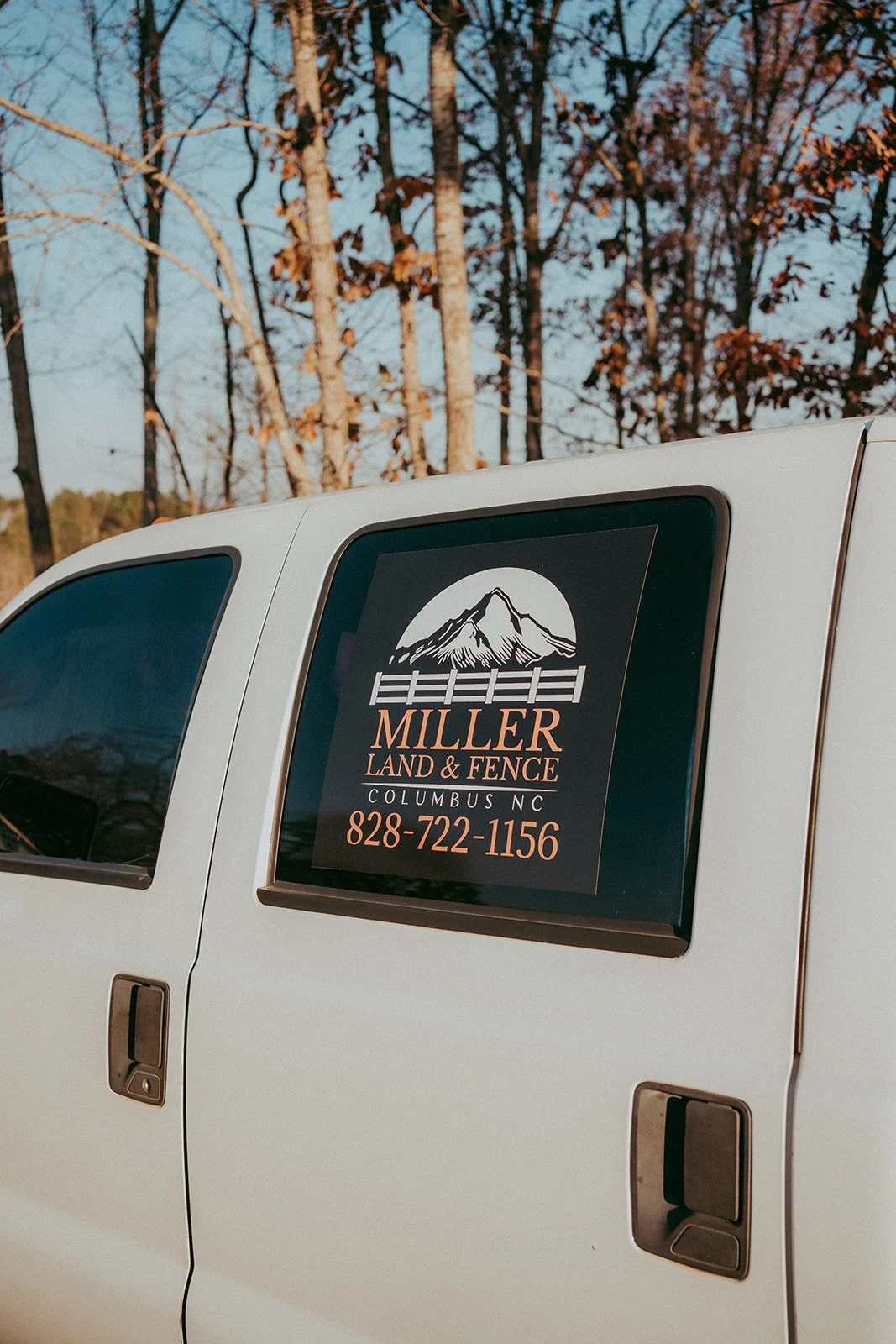 A white truck with a black and orange sign on the window that reads 'Miller Land & Fence, Columbus NC, 828-722-1156', featuring a mountain and fence graphic, parked outdoors during autumn with leafless trees in the background.