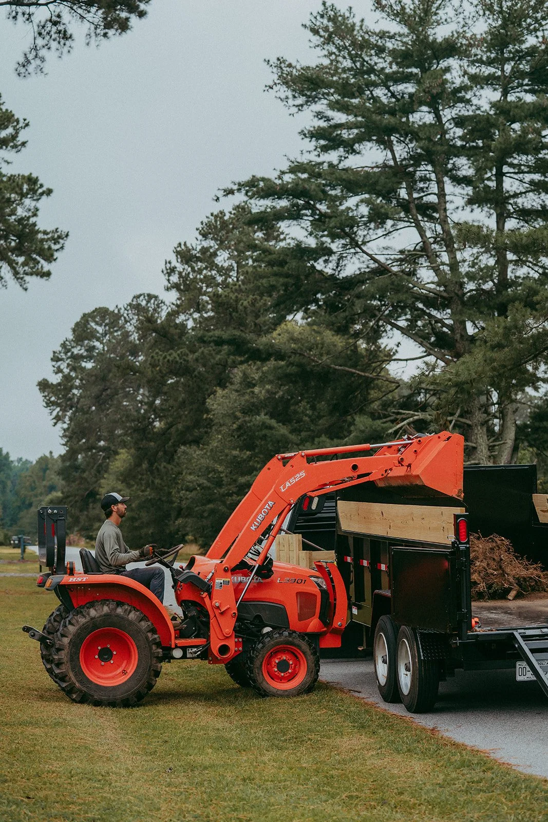 A man operating a Kubota tractor loader loaded with wood, parked next to a black dump trailer filled with mulch or wood debris, on a grassy area near trees.