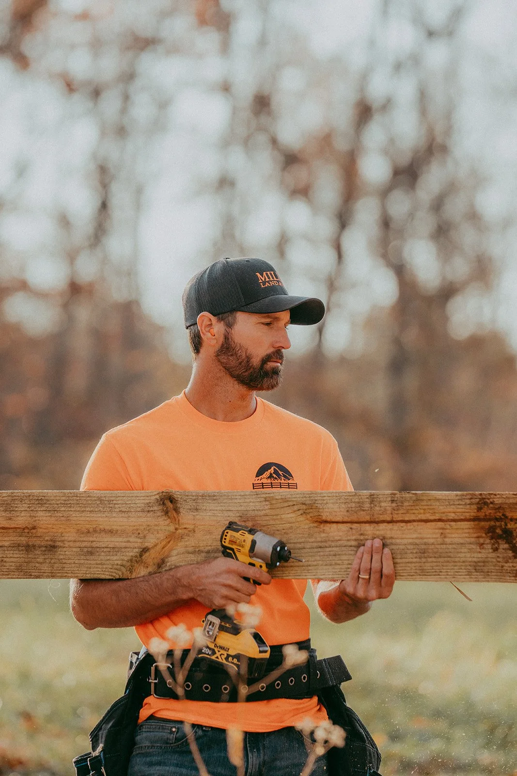 A man with a beard wearing an orange t-shirt and a black cap, holding a cordless drill and a piece of wood outdoors during daytime.