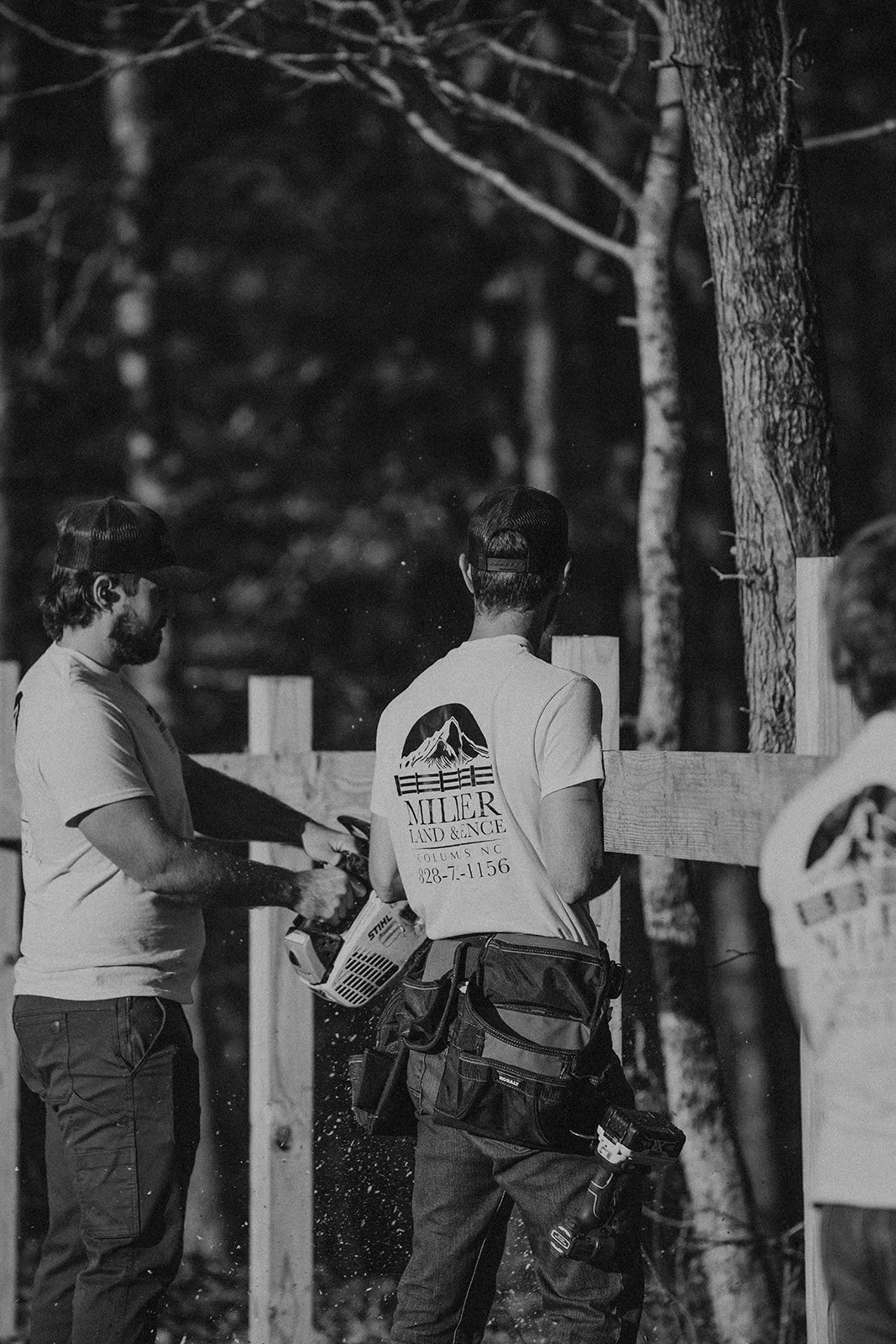 Three men working on a wooden fence, one with a saw and another with a tool belt, outdoors at night with trees in background.