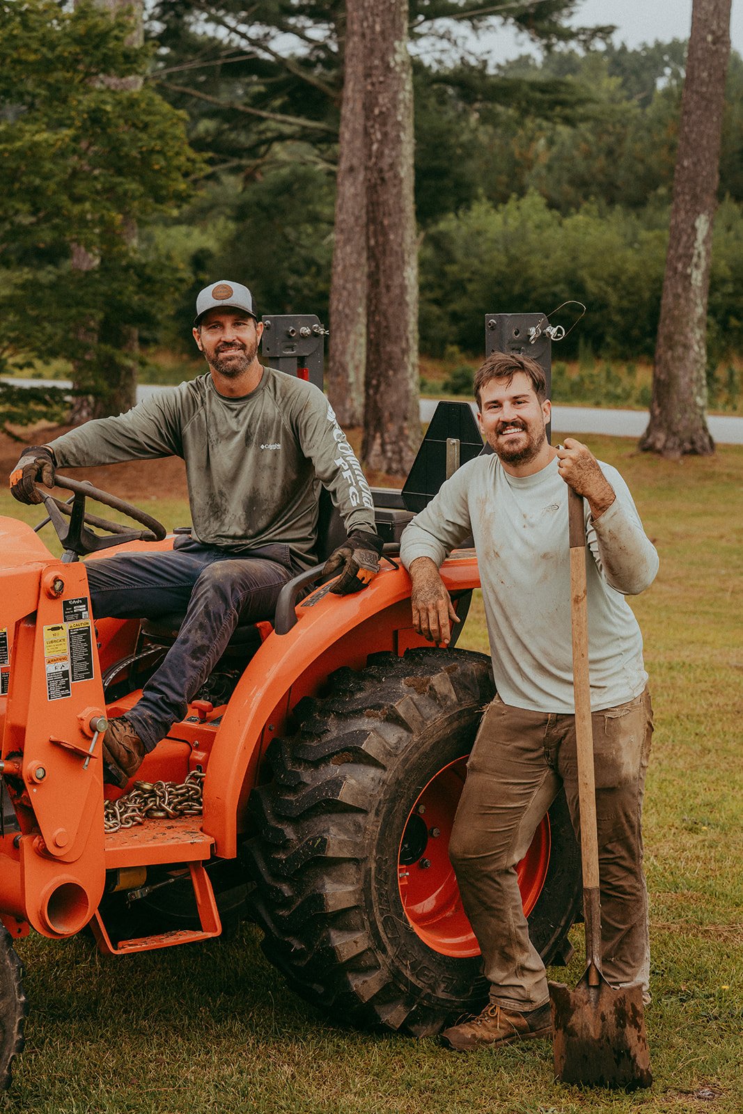 Two men with dirt-stained clothes posing with a tractor outdoors in a wooded area. One man is sitting on the tractor's seat, wearing a cap and gloves, while the other is standing beside it, holding a shovel and smiling.
