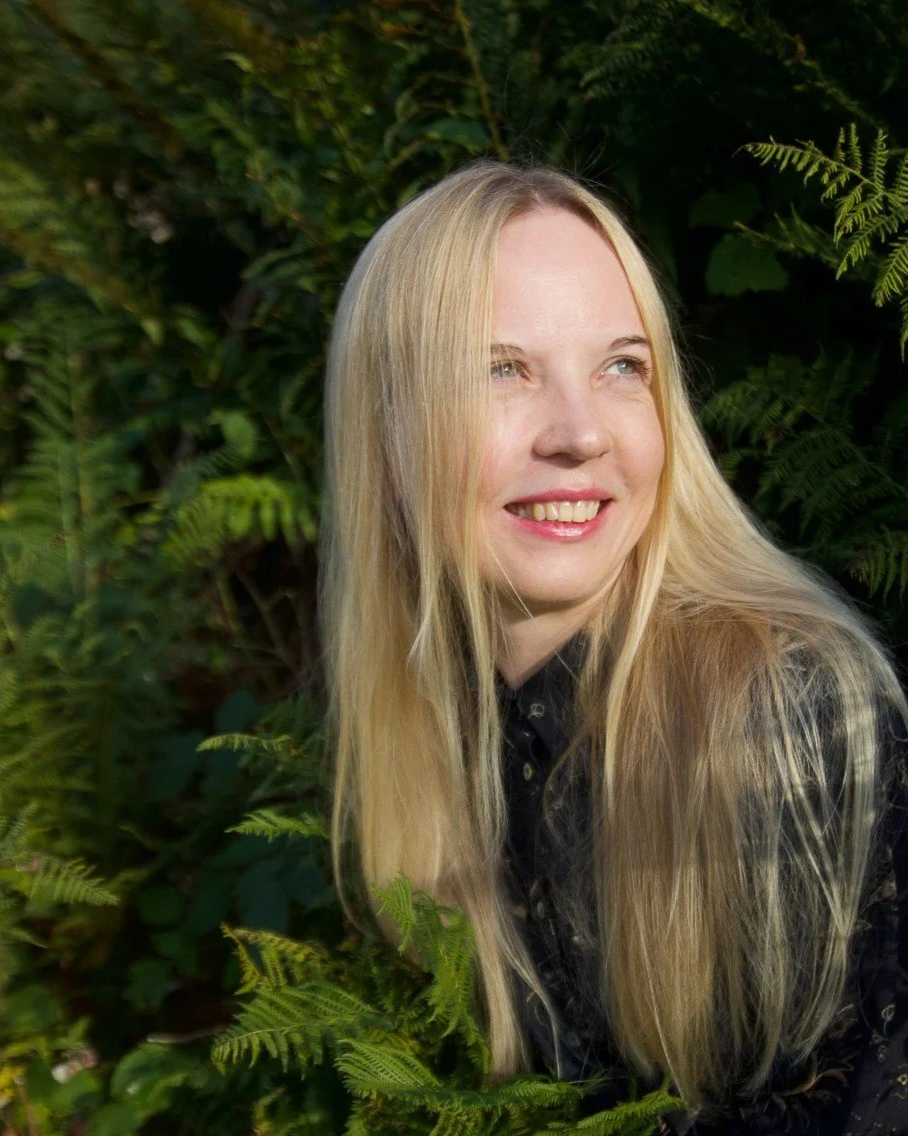 Junge Frau mit langen blonden Haaren lächelt und blickt nach rechts, umgeben von grünen Blättern und Fichtenzweigen in der Natur.