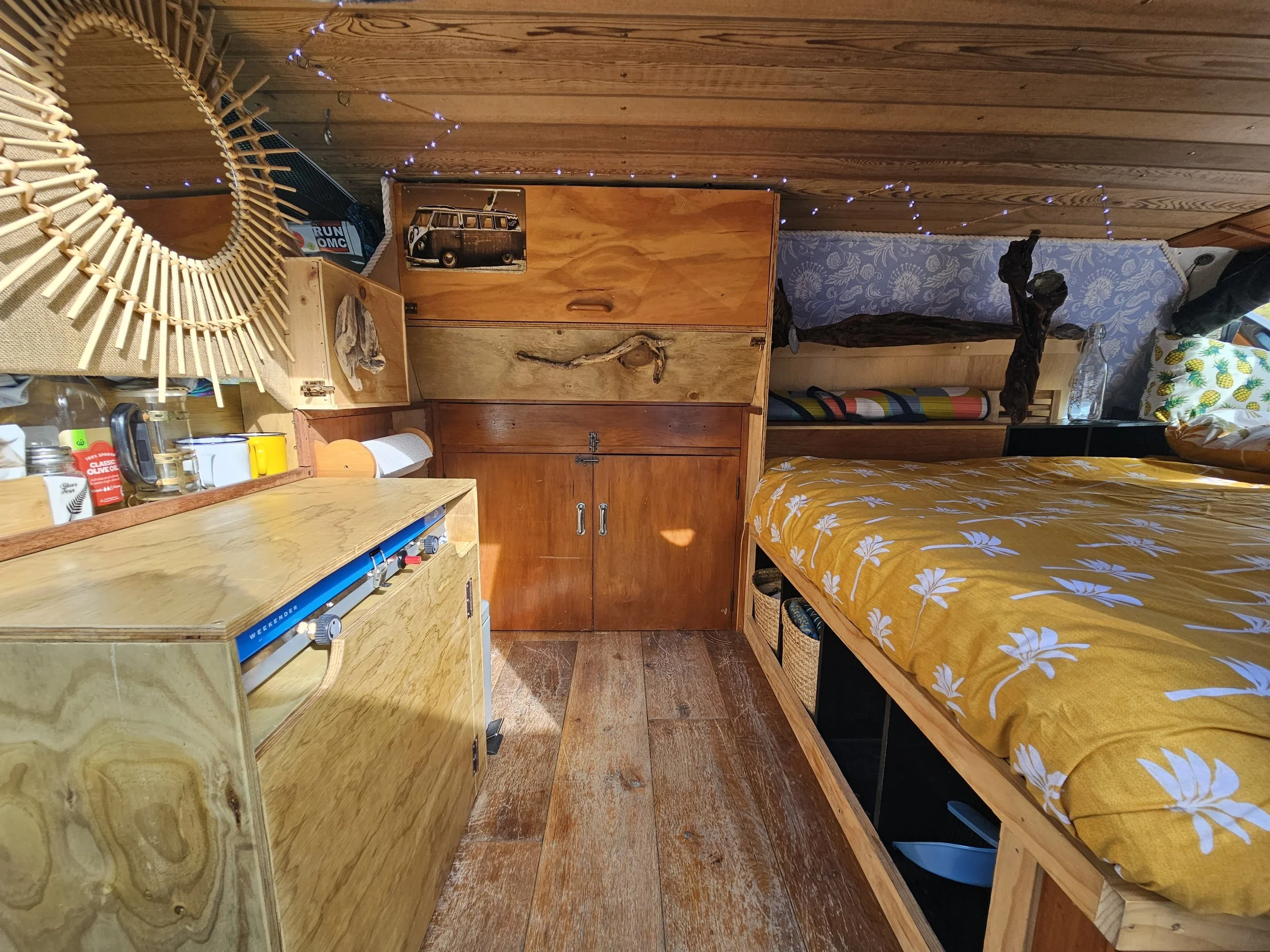 A cozy bedroom with wooden furniture, including a wardrobe, a bed with yellow and white floral bedding, and a wooden cabinet. There are decorative string lights hanging across the ceiling, a mirror with a wicker frame, and various items like a framed picture of a vintage van, and some colorful pillows.