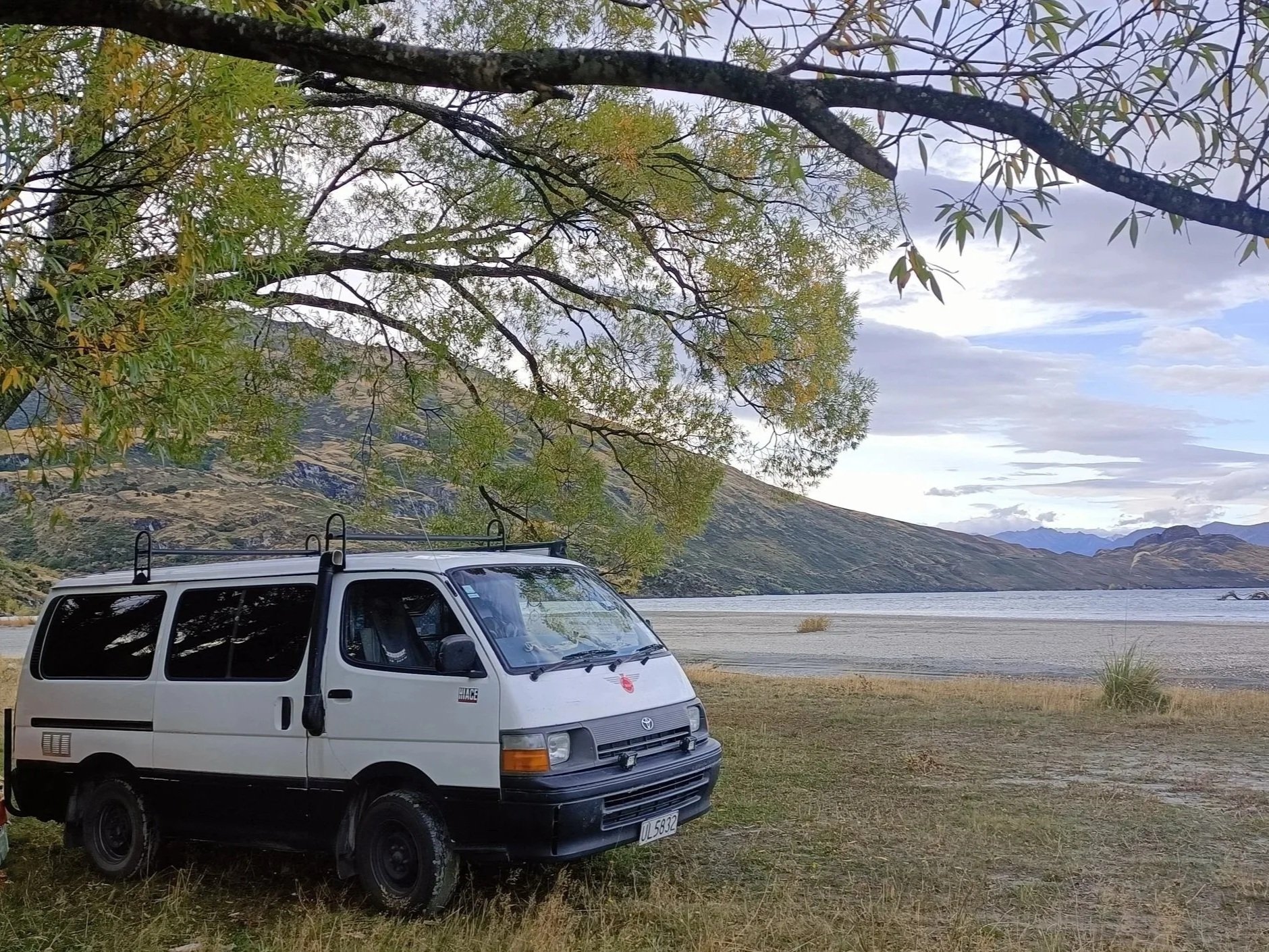 A white Toyota HiAce van parked on grass near a lake Wanaka, with mountains in the background and trees overhead.