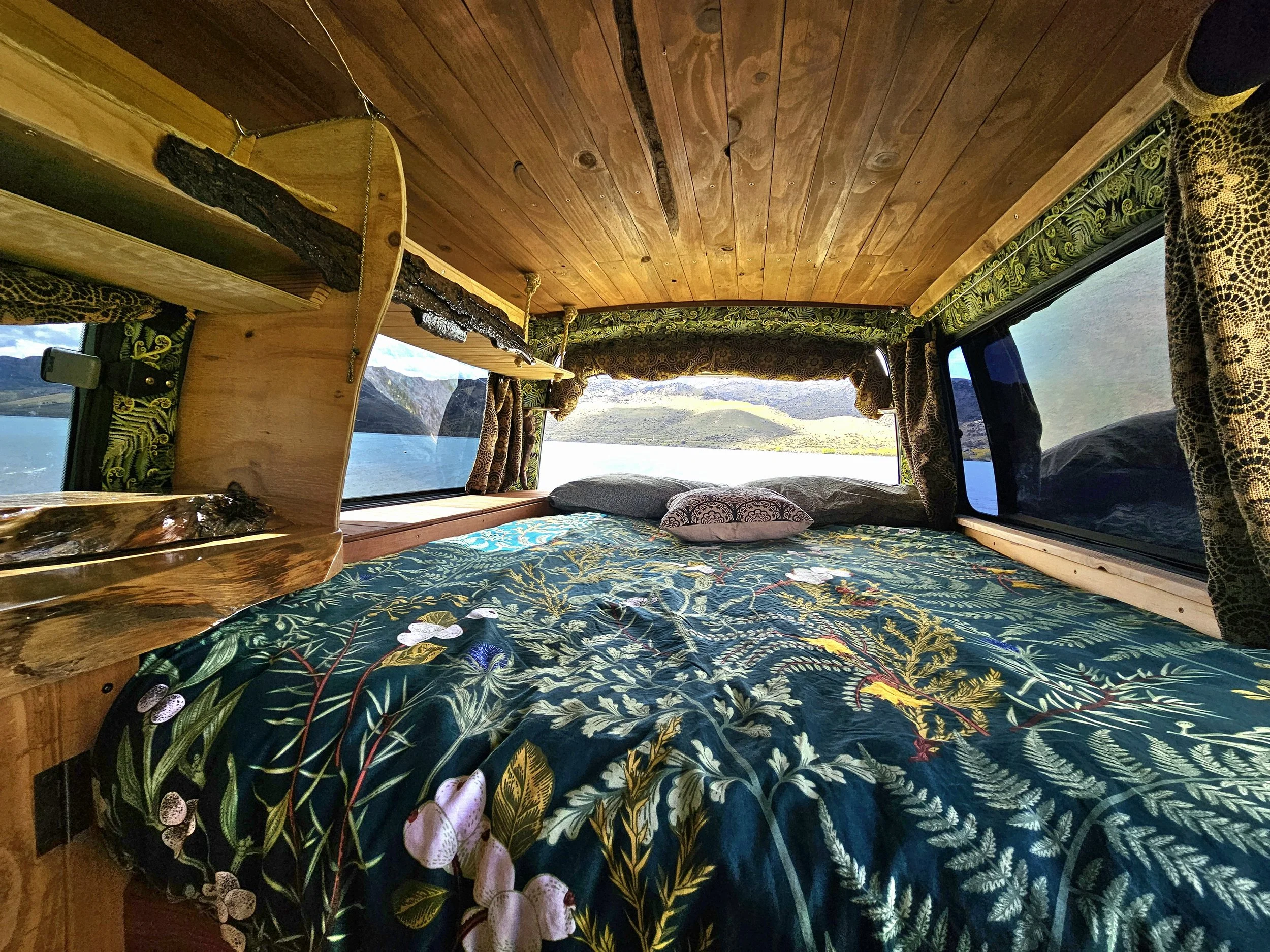 Interior of a wooden camper van with a bed covered in a kiwiana print bedspread, large windows showing a scenic lake and mountains, and decorative curtains.