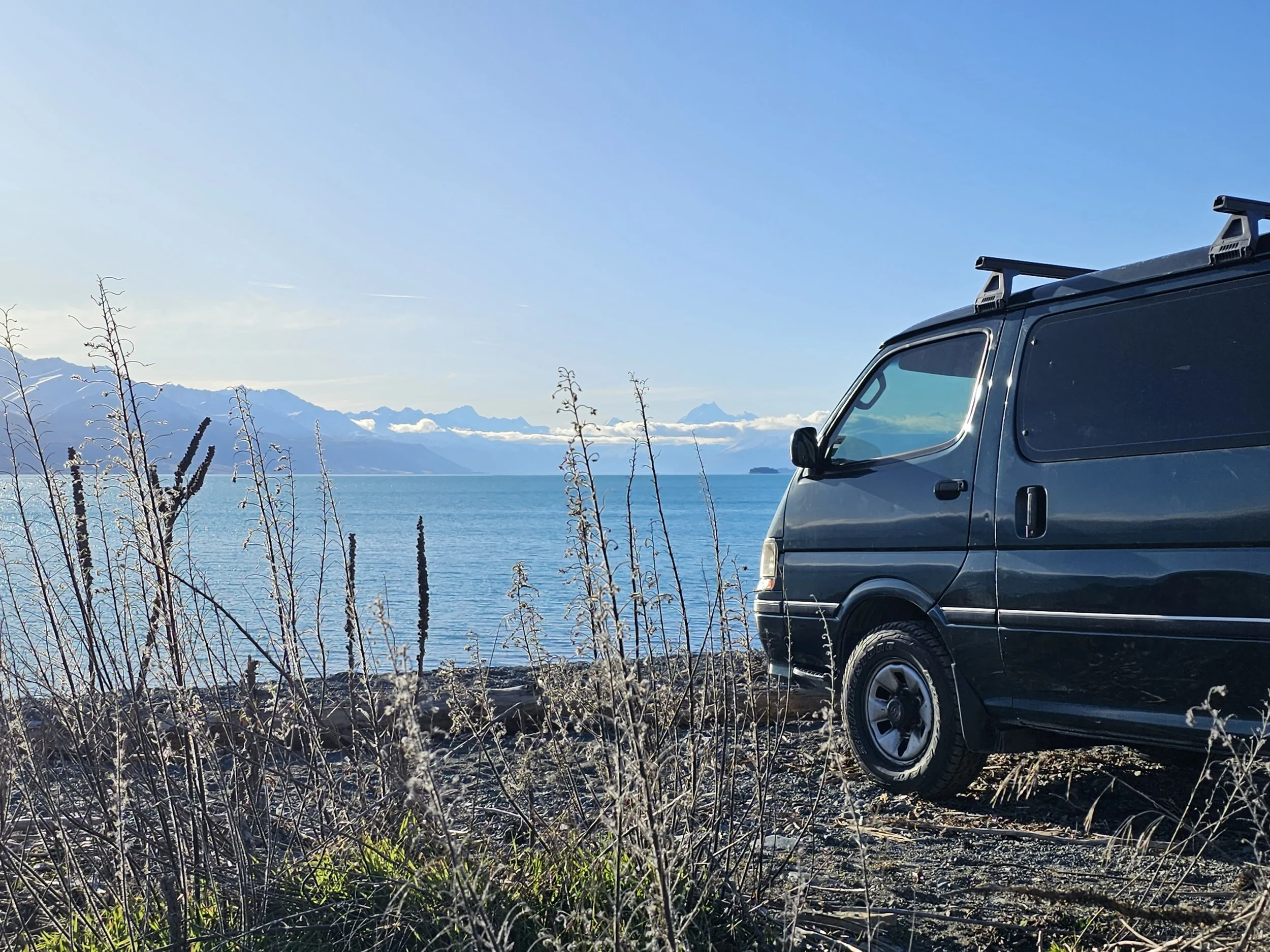 A black van parked near Lake Pukaki with Mount Cook in the background, some dry plants in the foreground, and a clear blue sky.