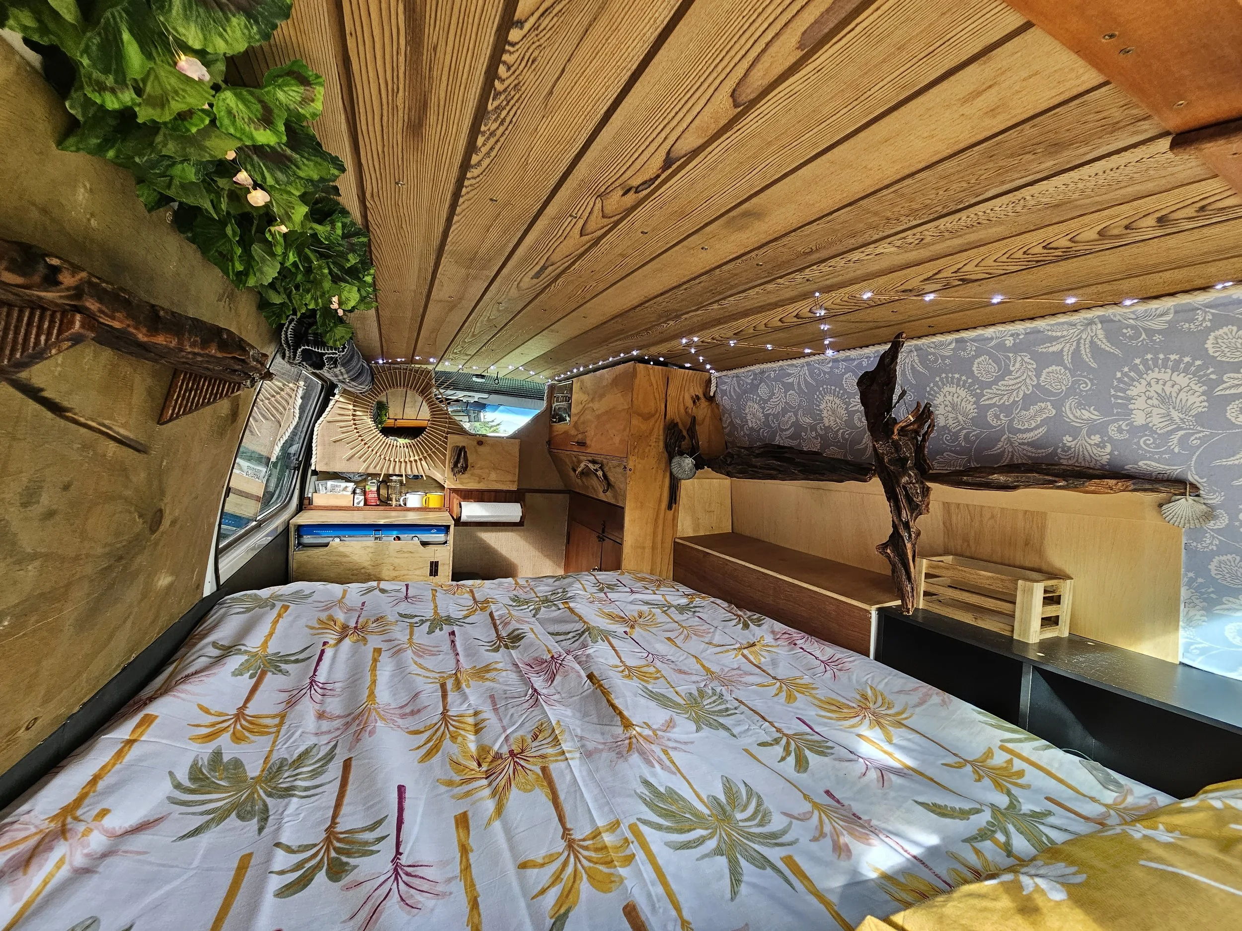 A cozy room with a bed covered in a floral yellow, green, and white bedspread, a wooden desk, leafy green plants on a driftwood ledge, and fairy lights on a wooden ceiling.