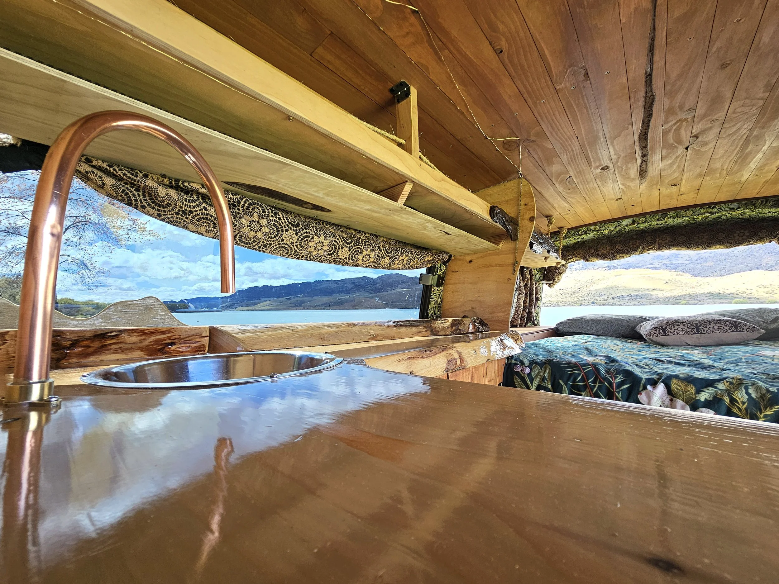 Interior of a wooden camper with a view of a lake and mountains outside the window, featuring a countertop with a copper sink, patterned curtains, and a double bed.