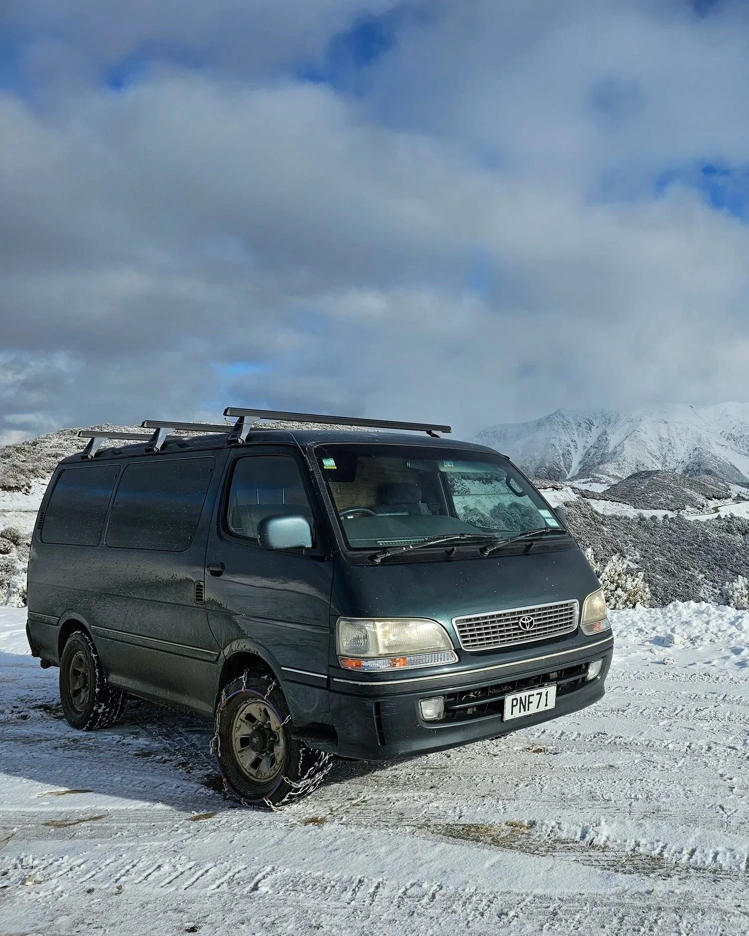 A black Toyota van with snow tires parked in a snowy landscape with snow-covered mountains and cloudy sky in the background.