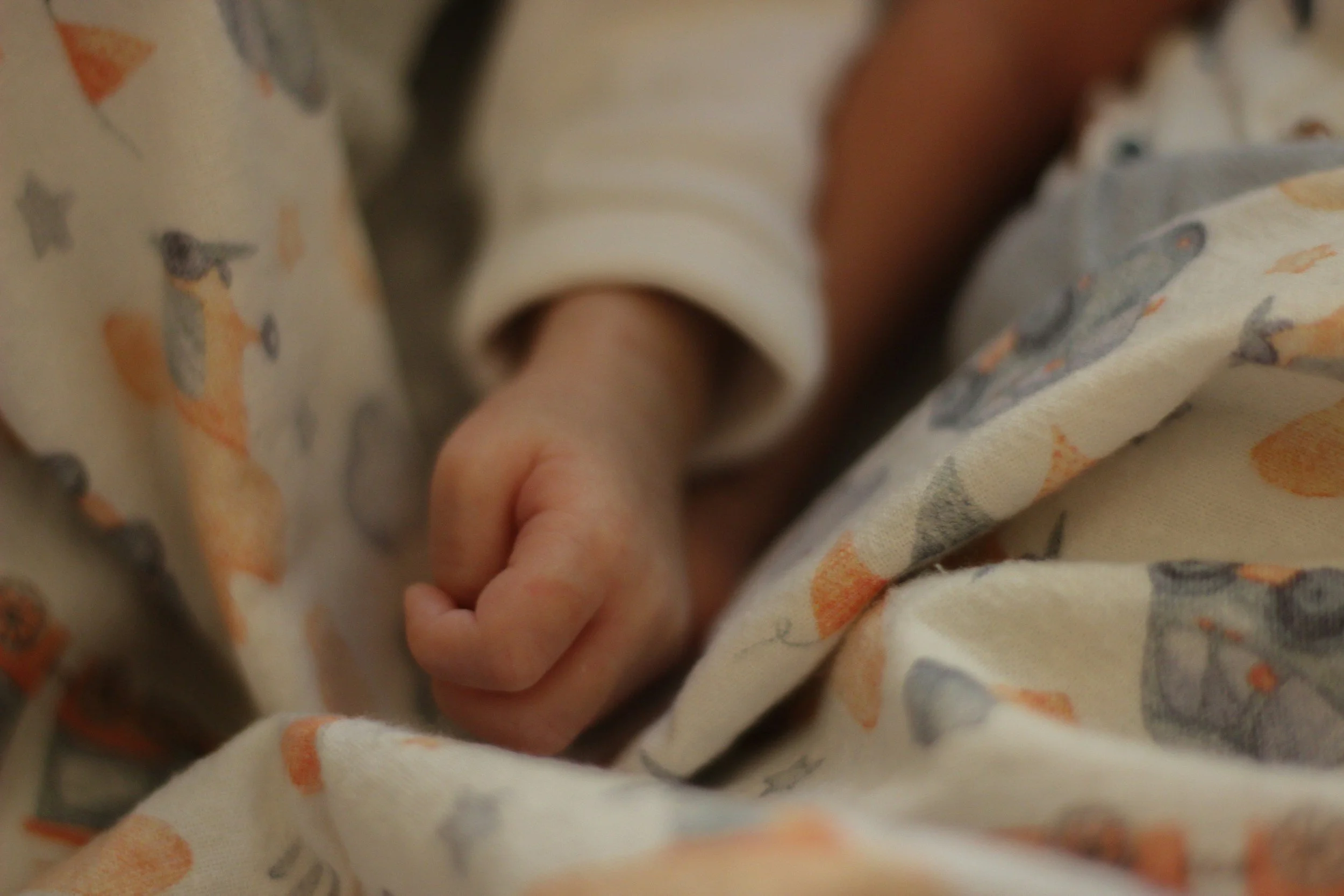 Close-up of a baby's hand clenched into a fist, resting on patterned fabric with small animal images, wearing a white long-sleeve shirt.