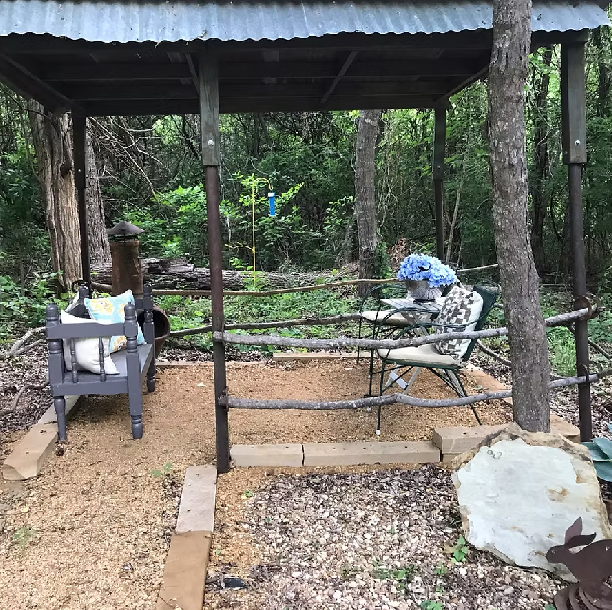 A cozy outdoor seating area under a rustic wooden shelter in a wooded backyard, with a small table holding a blue flower arrangement and two chairs with cushions, surrounded by a simple wooden fence and natural greenery.