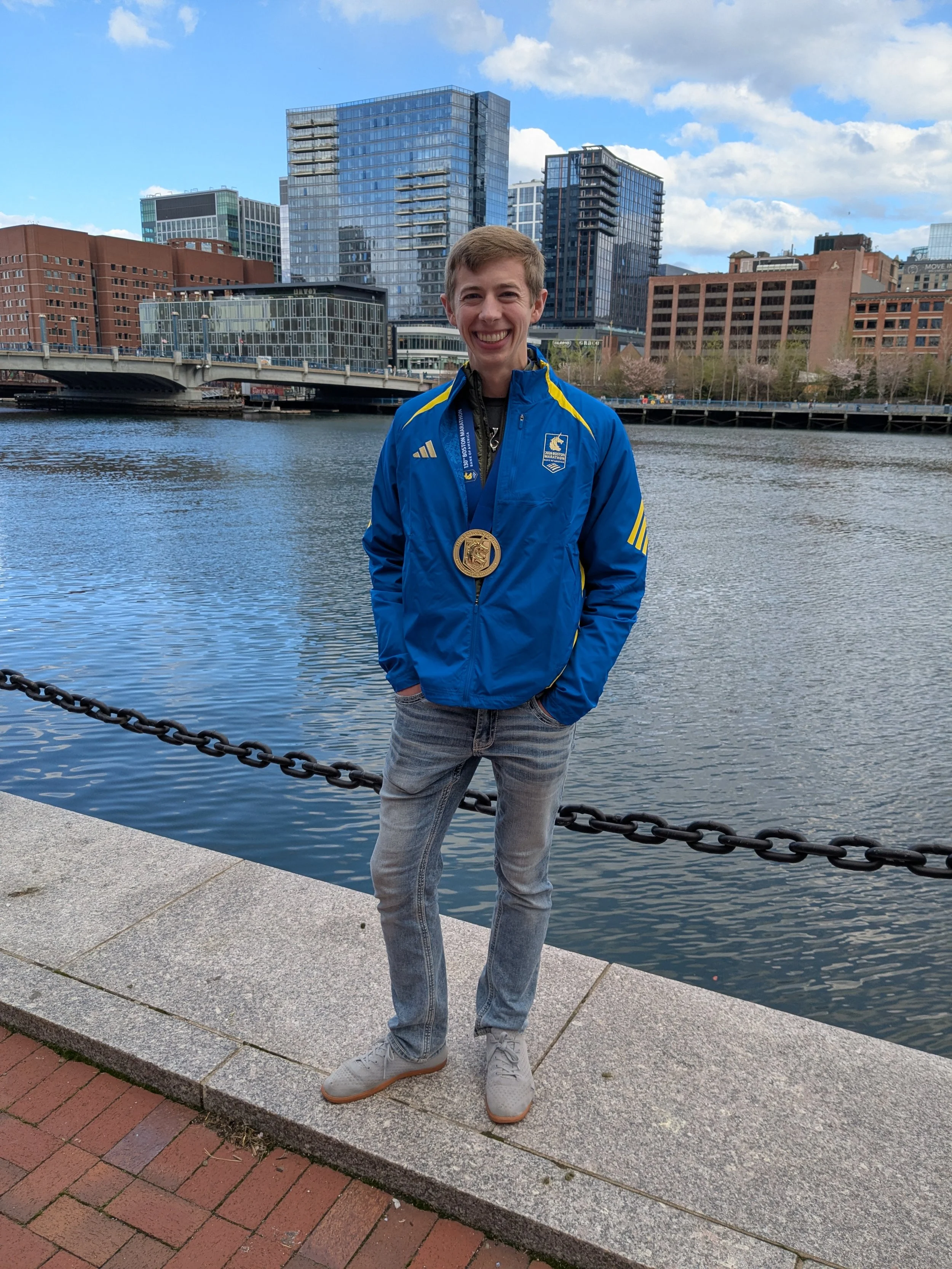 A smiling young man with short hair wearing a blue Adidas jacket with a medal around his neck, standing on a concrete walkway by a waterway with a city skyline of modern glass buildings in the background.