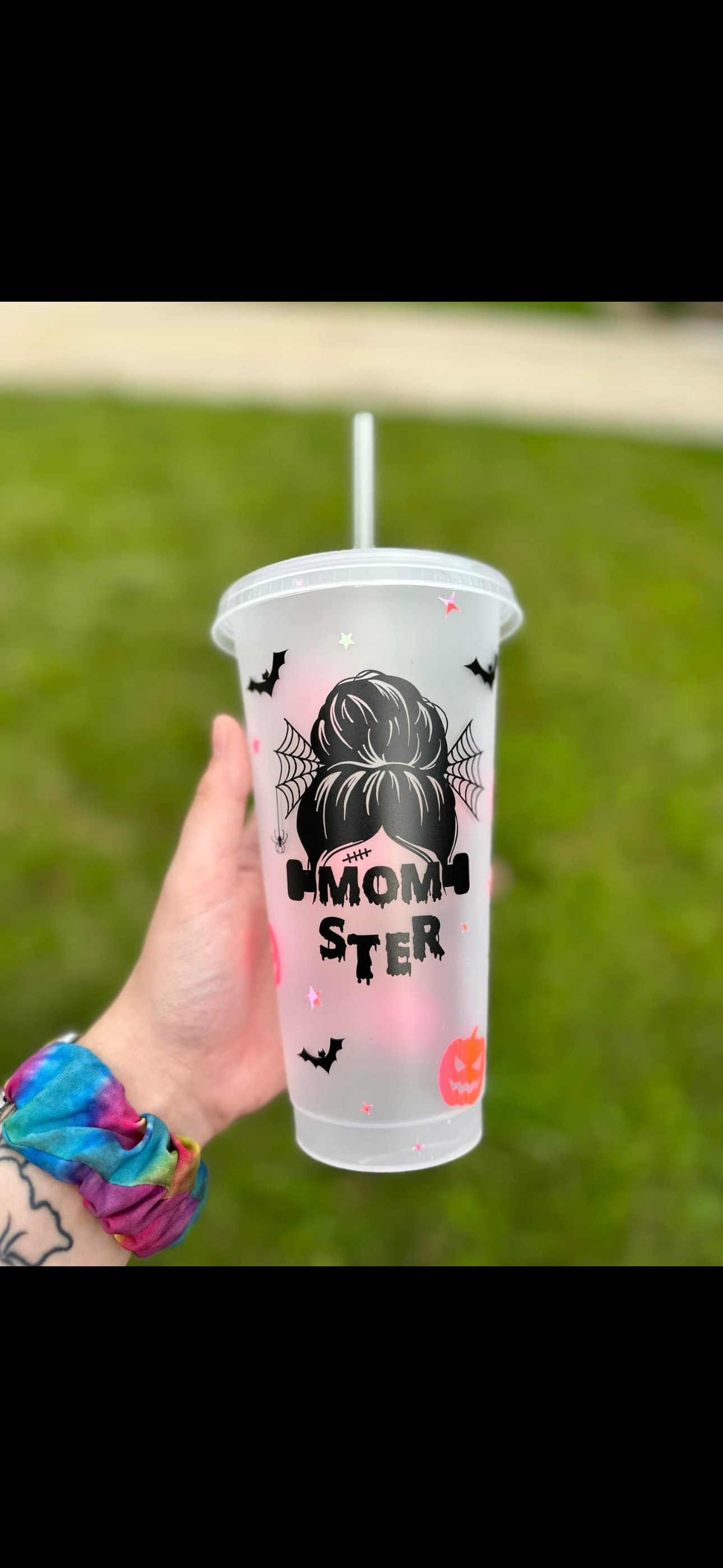 Hand holding a clear plastic cup with Halloween themed design, including a silhouette of a girl with a bun hairstyle, spider webs, bats, a smiling pumpkin, and the words 'Cool Mom' on it, with a straw.