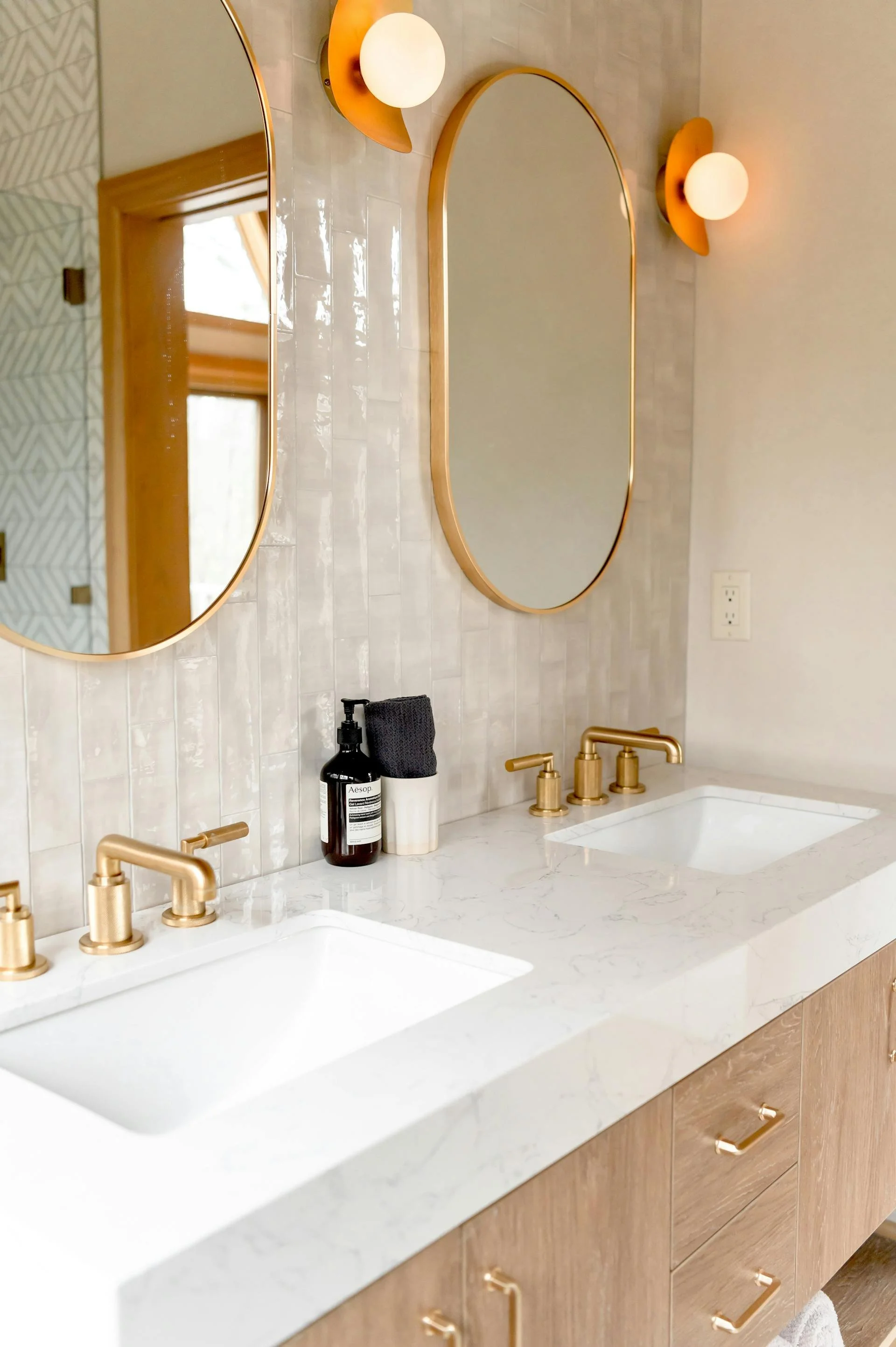 A modern bathroom vanity with a white marble countertop, two rectangular sinks, gold fixtures, large oval mirrors, and two wall-mounted spherical light fixtures. There is a black soap bottle and towel on the counter.
