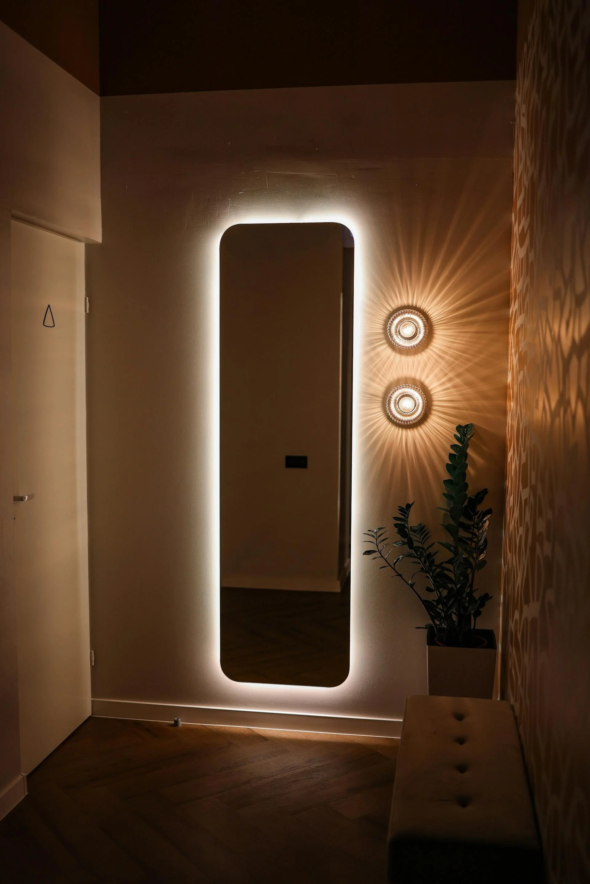 A hallway with a tall, rectangular mirror on the wall. The mirror is backlit with LED lighting, creating a soft glow around its edges. Two decorative wall sconces with intricate designs cast starburst patterns of light and shadow on the wall beside t