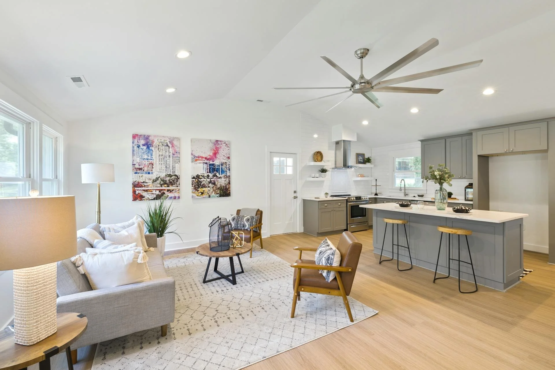 Open-concept living room and kitchen with white walls, light wood floors, gray cabinets, and modern furniture, including a beige sofa, wooden armchair, and a kitchen island with barstools.