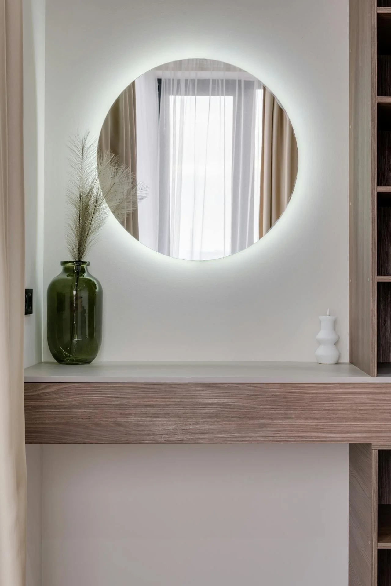 Decorative interior mirror with backlit illumination, a green vase with dried grass, and a small white vase on a wooden shelf.