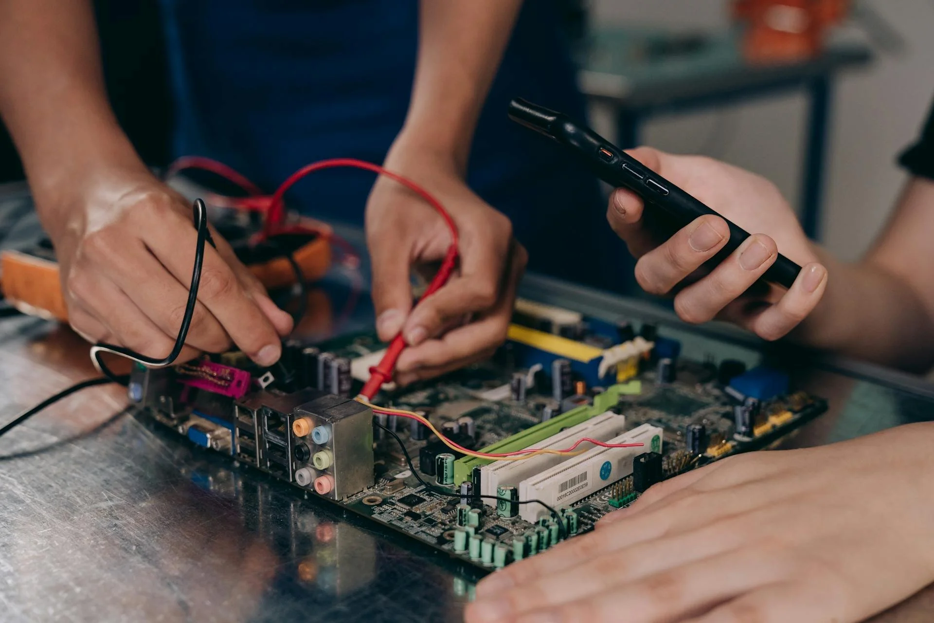 Two people working on a computer motherboard, one holding a smartphone and the other adjusting connections with tools, on a metal workbench.