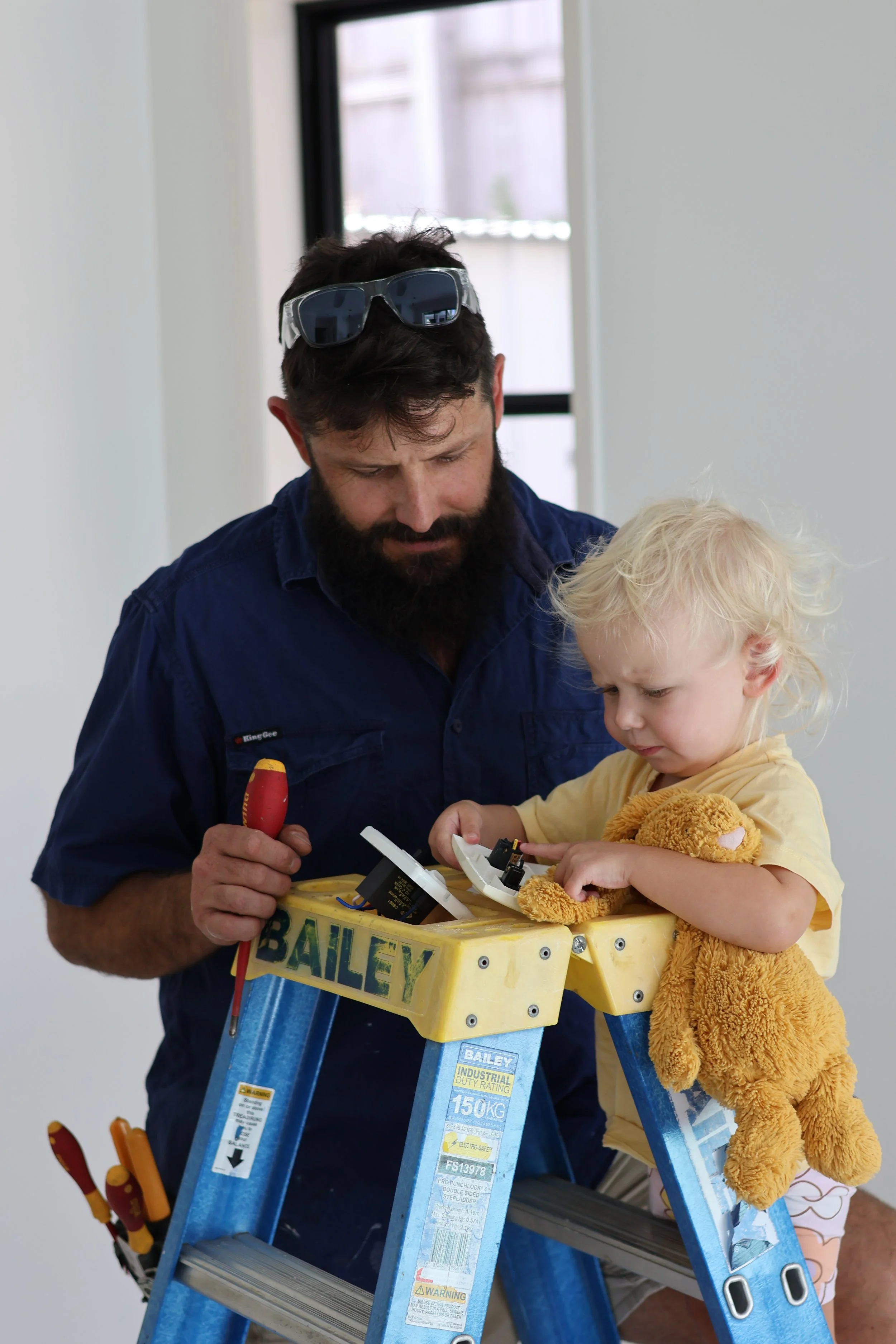 A man and a young girl are working on an electrical project together on a yellow ladder, with the man holding a screwdriver and the girl examining the wiring.