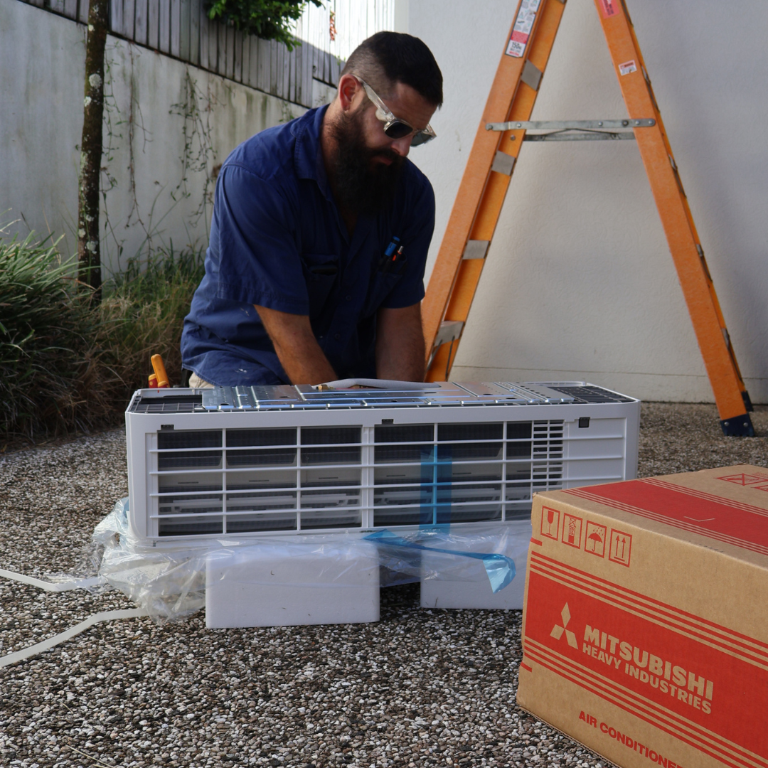 A man in blue work clothes and sunglasses installs an air conditioner outdoors, with a ladder and a cardboard box nearby.