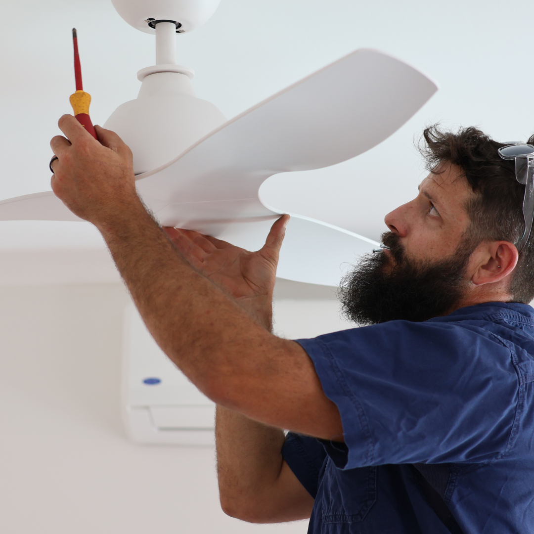 A man with a beard wearing glasses on his head, inspecting a white ceiling fan while holding a screwdriver, in a room with white walls and air conditioning unit.