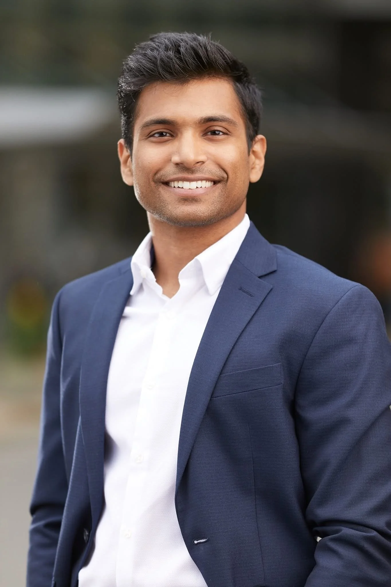 A young man with dark hair and a smile, wearing a navy blazer and white shirt, standing outdoors with blurred background.