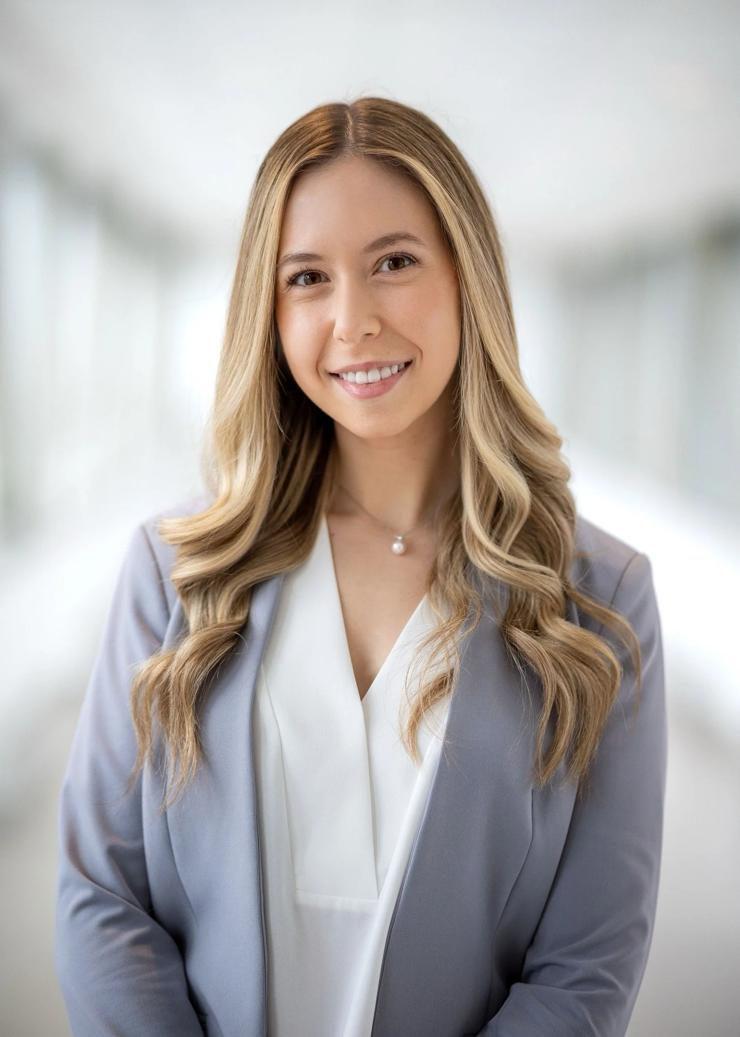 Professional woman with long wavy blonde hair smiling, wearing a white blouse and a light gray blazer, in an indoor office setting.