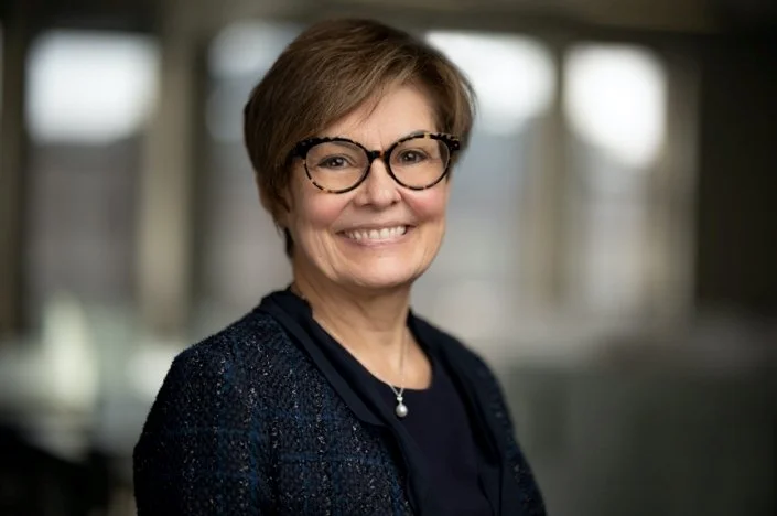 A smiling middle-aged woman with short brown hair, glasses, and a dark blue jacket, standing indoors with large windows in the background.