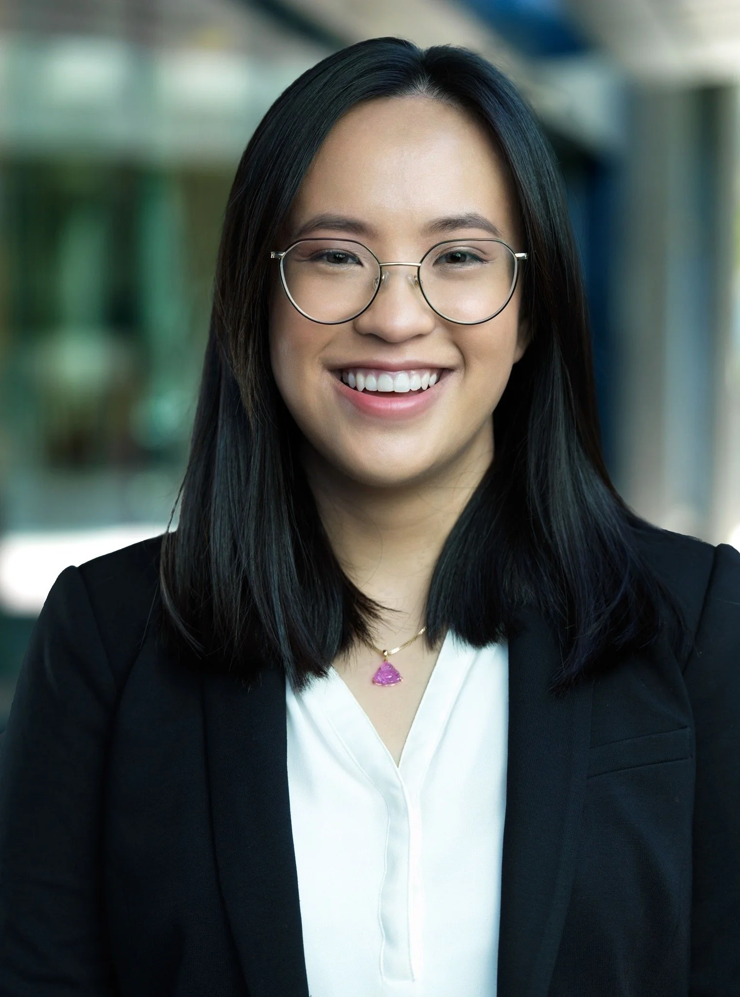 A young woman smiling, wearing glasses, a white blouse, and a black blazer, with a blurred background.