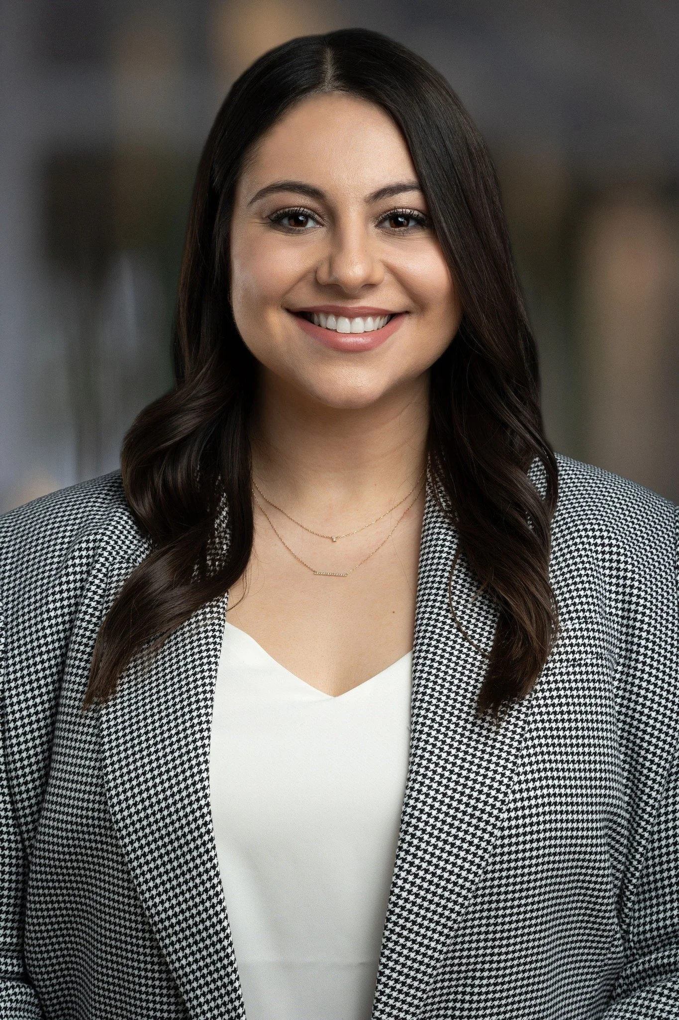 Portrait of a young woman with dark brown hair in loose waves, smiling, wearing a houndstooth blazer over a white top and layered delicate gold necklaces.