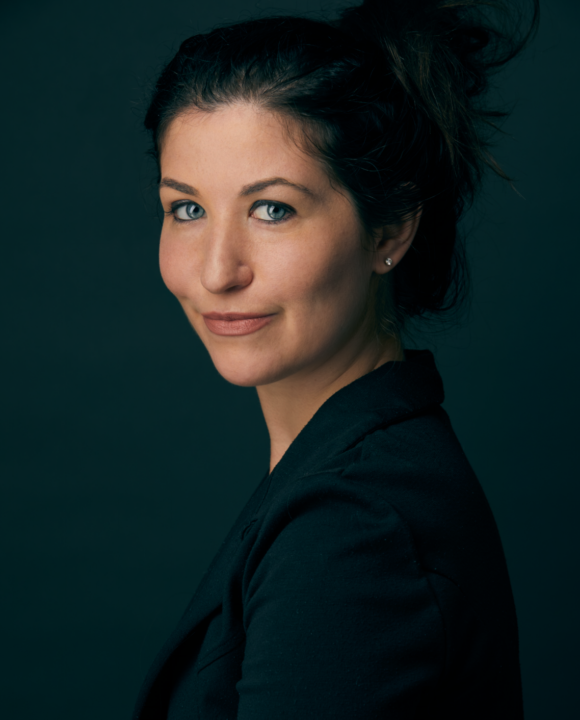 Portrait of a woman with light skin, blue eyes, and dark brown hair styled in a loose updo, wearing a black blazer against a dark background.