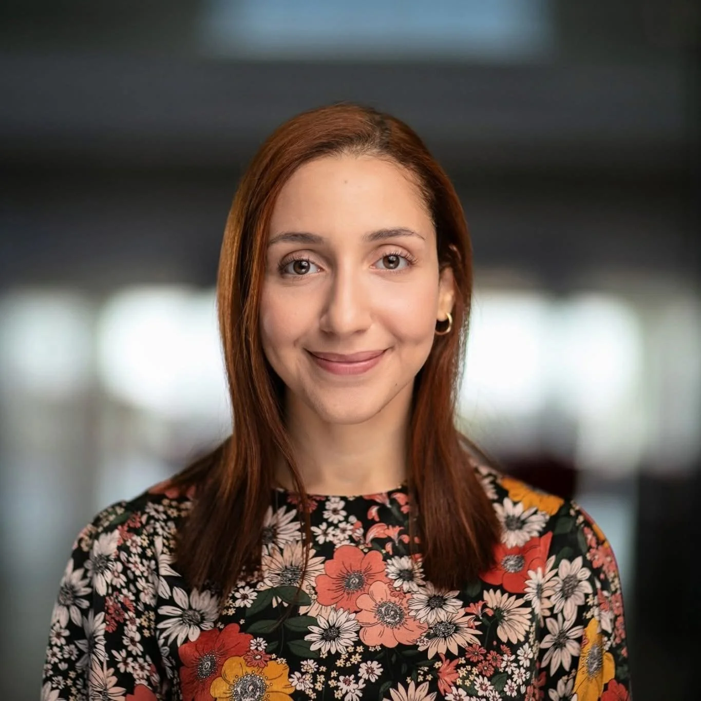Close-up of a young woman with reddish-brown hair wearing a floral dress, smiling, in an indoor setting with blurred background.
