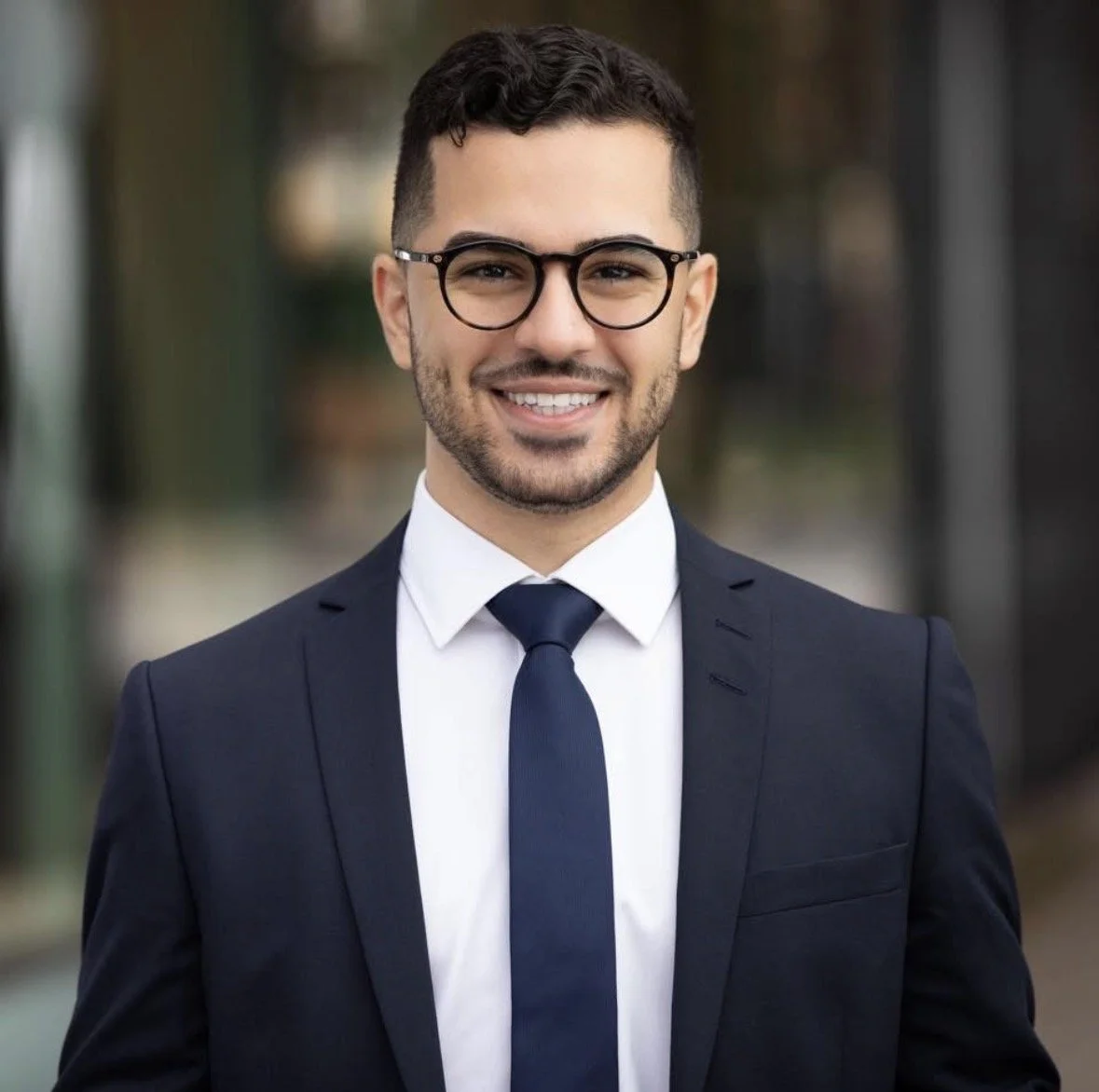 A young man with dark hair, glasses, and a beard, wearing a formal navy suit, white shirt, and navy tie, smiling at the camera outdoors.