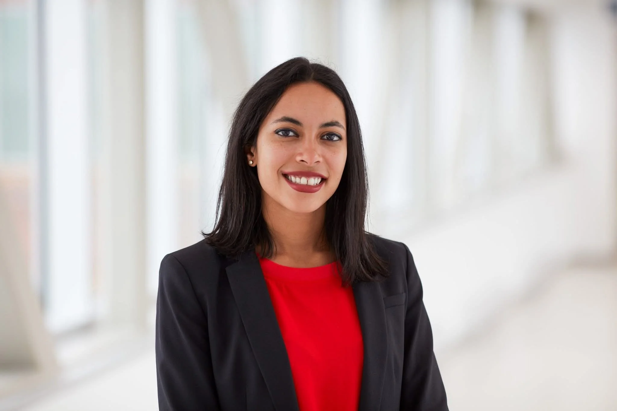 A young woman with black hair in a professional blazer and red top smiling in an indoor setting with bright, blurred background.