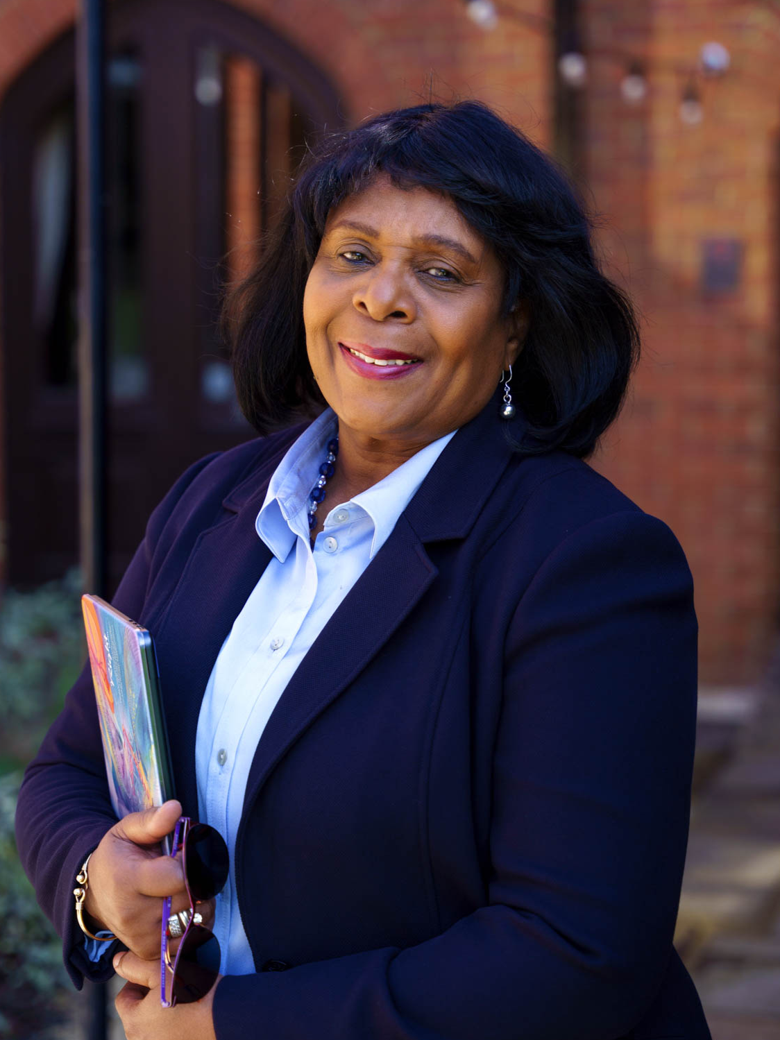 A woman in a dark blazer and white shirt holding a colorful folder and glasses, standing outside near a brick building, smiling at the camera.