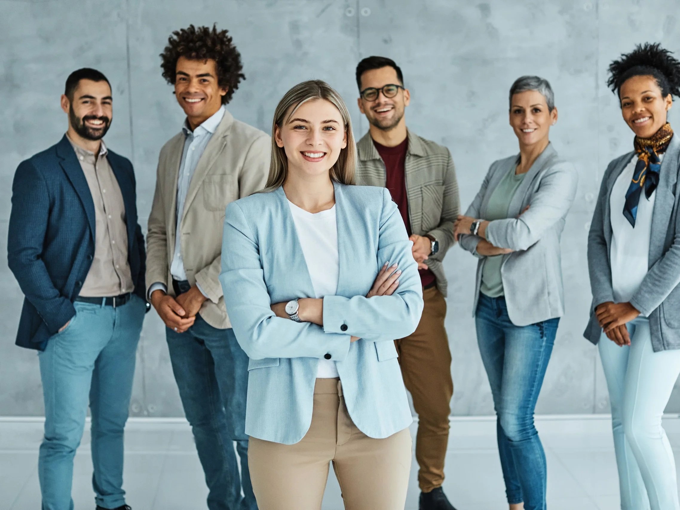 A diverse group of six professionals standing together in a modern office, smiling at the camera, with a woman in a light blue blazer in the foreground.