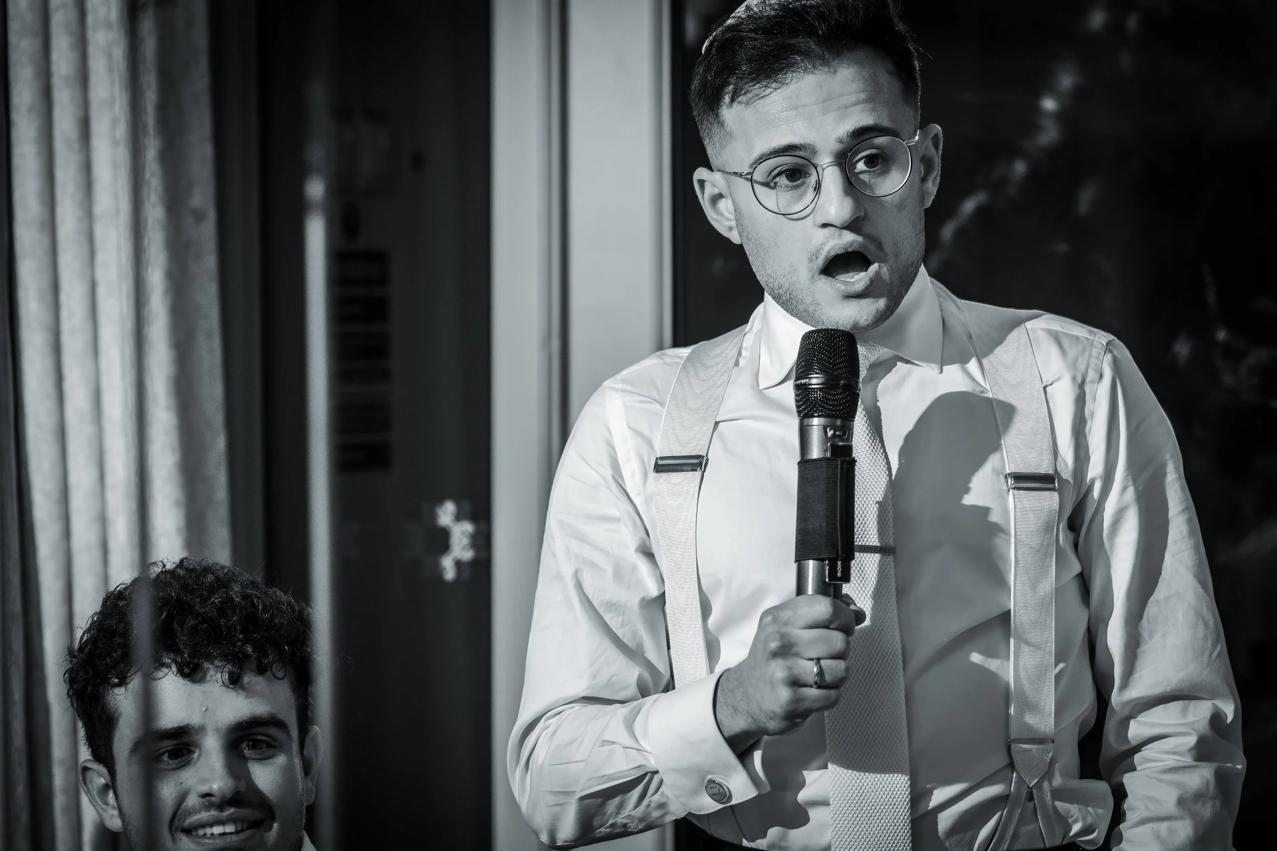 A man wearing glasses, a white shirt, suspenders, and a tie is speaking into a microphone. A partial face and head of another person are visible in the foreground, smiling. The scene appears to be indoors near a window.