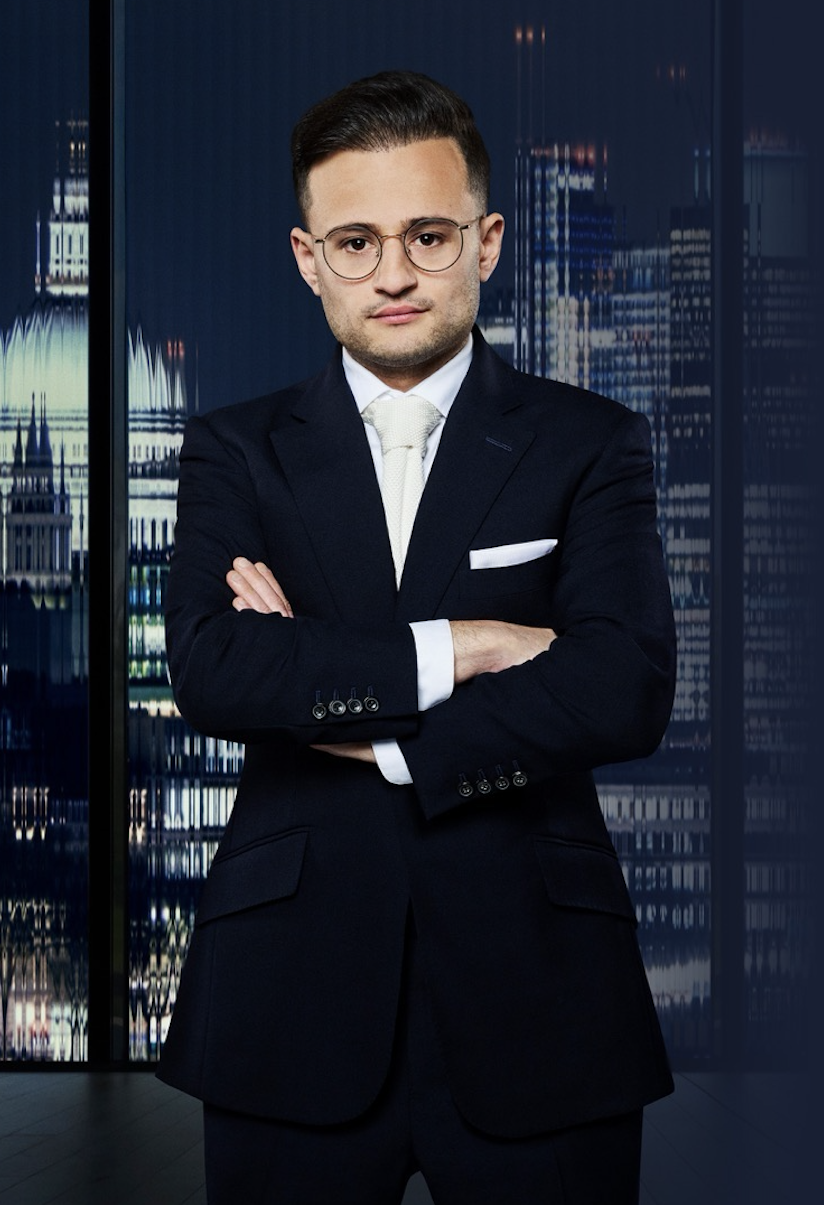 A young man in a dark suit, white shirt, and white tie standing with arms crossed in front of a cityscape view through large windows.