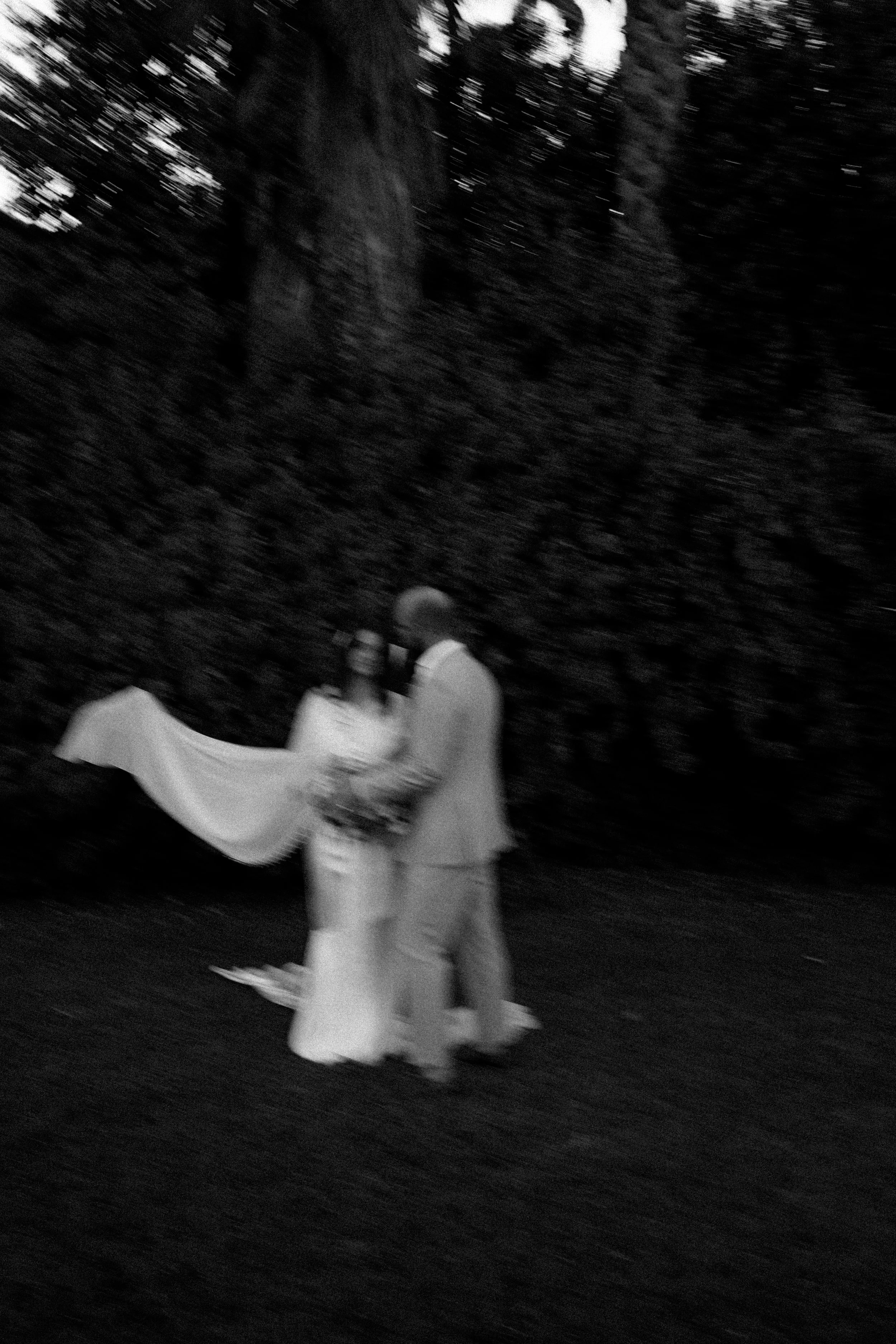 A black and white photo of a couple holding hands and exchanging rings outdoors near trees, during a wedding ceremony.
