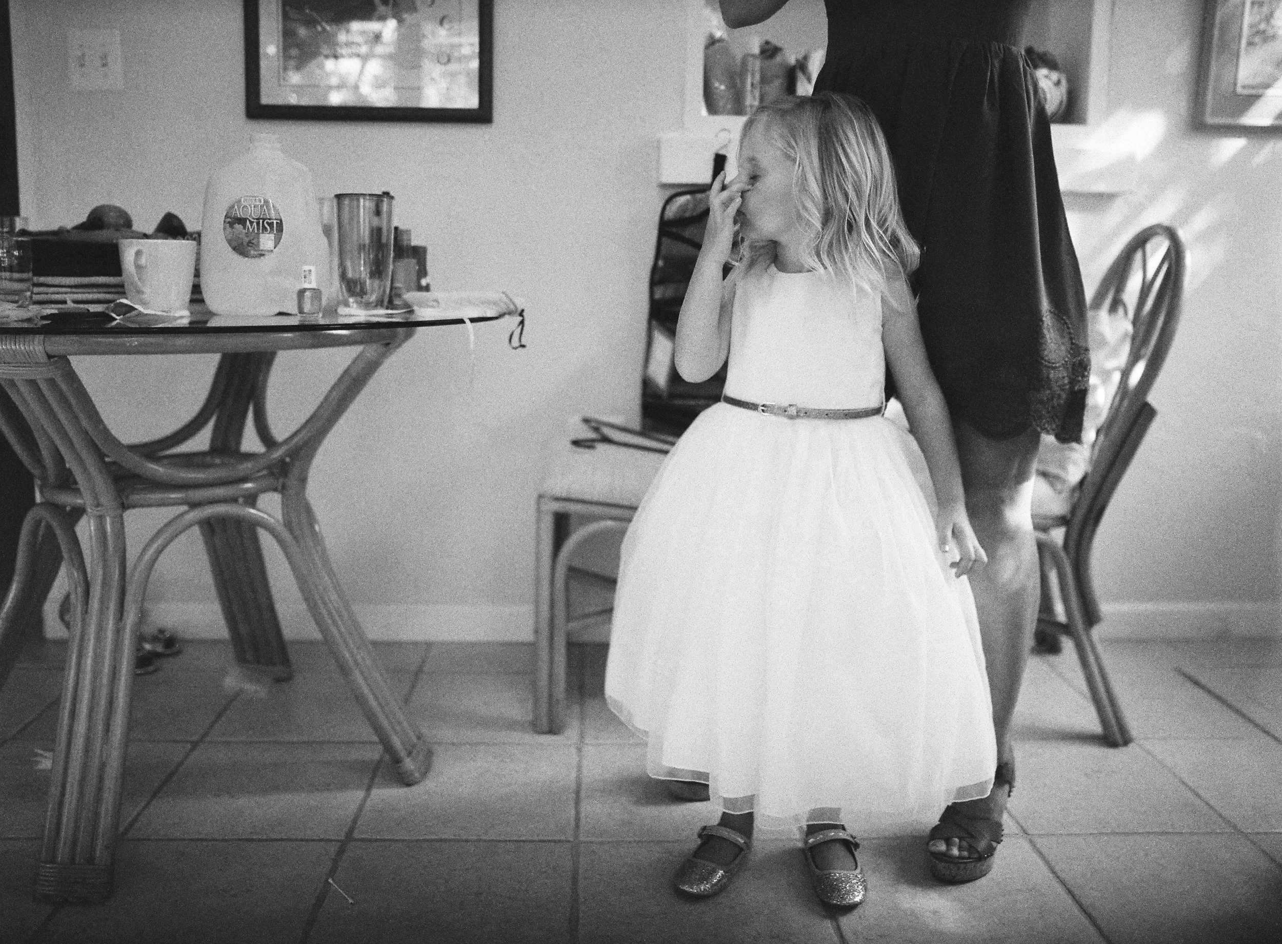 A young girl in a white dress with a belt, wearing glittery shoes, standing indoors, touching her face with one hand. She's beside an adult woman whose face is not visible, and she's wearing a dark outfit. The background has a table with various item