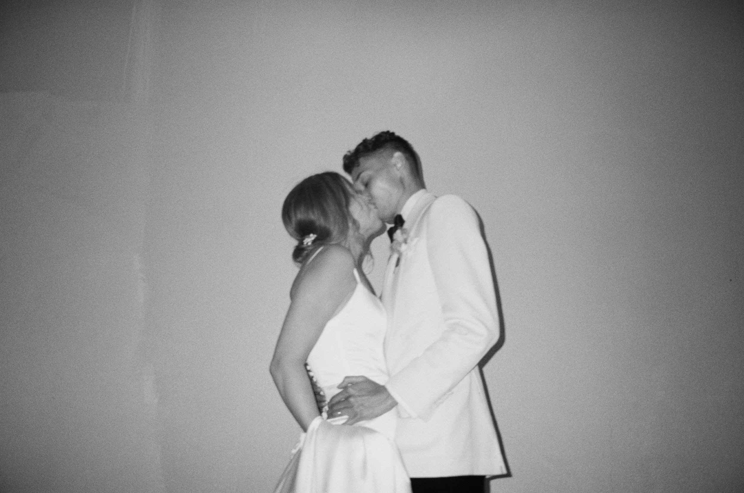 A black-and-white photo of a bride and groom sharing a kiss, with the groom in a white tuxedo and the bride in a white wedding dress, against a plain background.